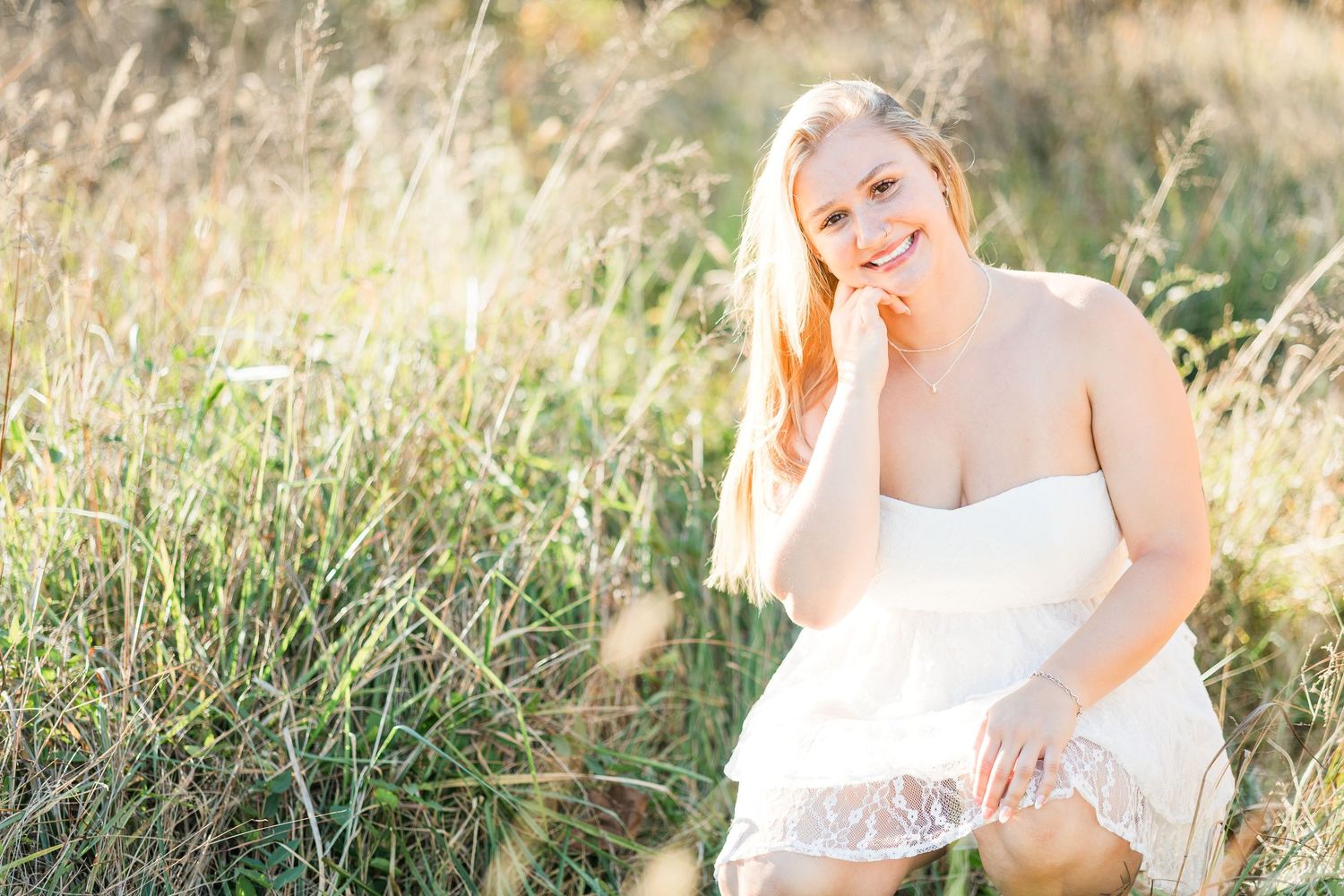A person in white dress poses in a sunny field with tall grass.