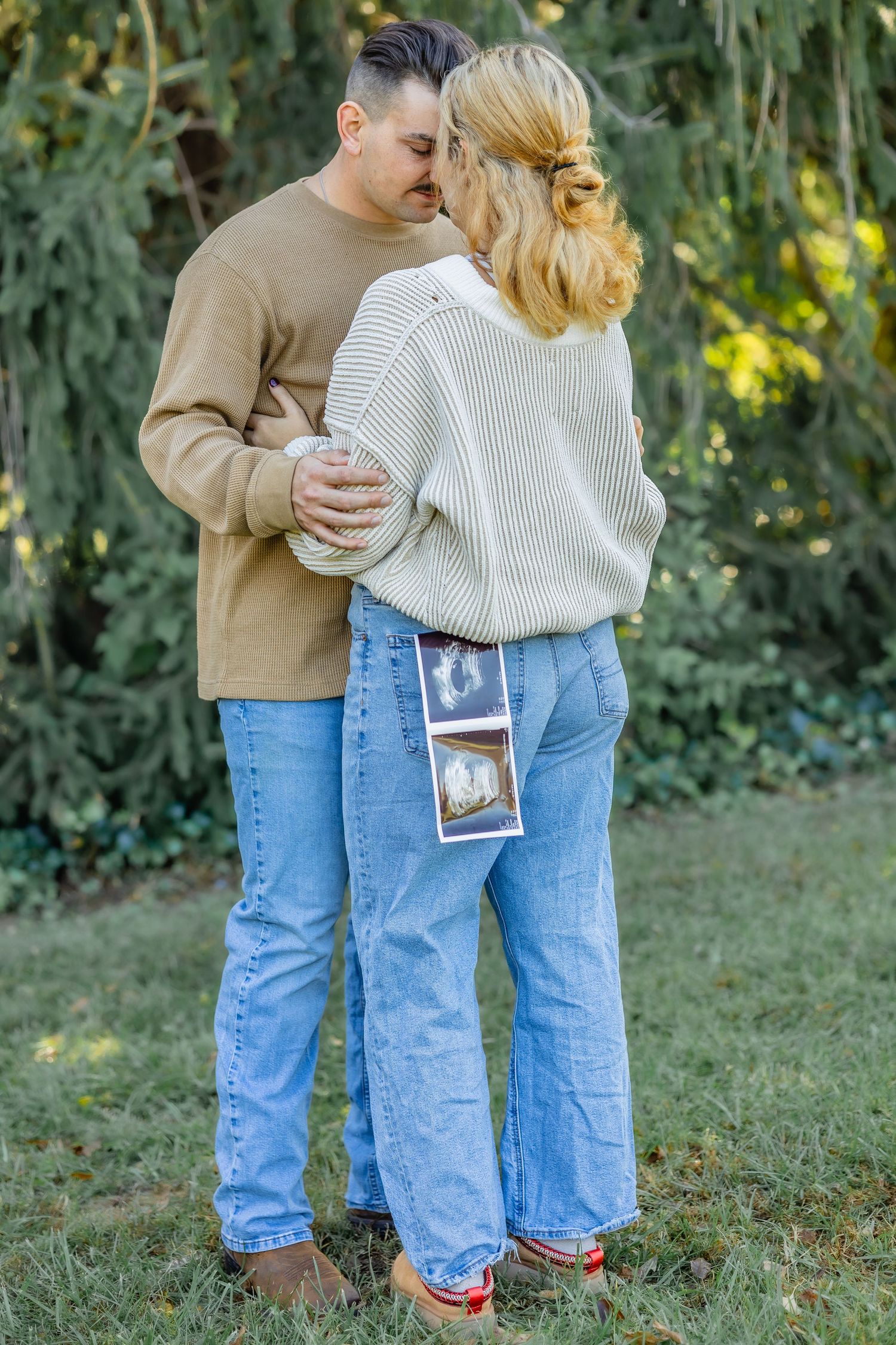 Intimate moment between couple with sonogram photos in pocket against green foliage.