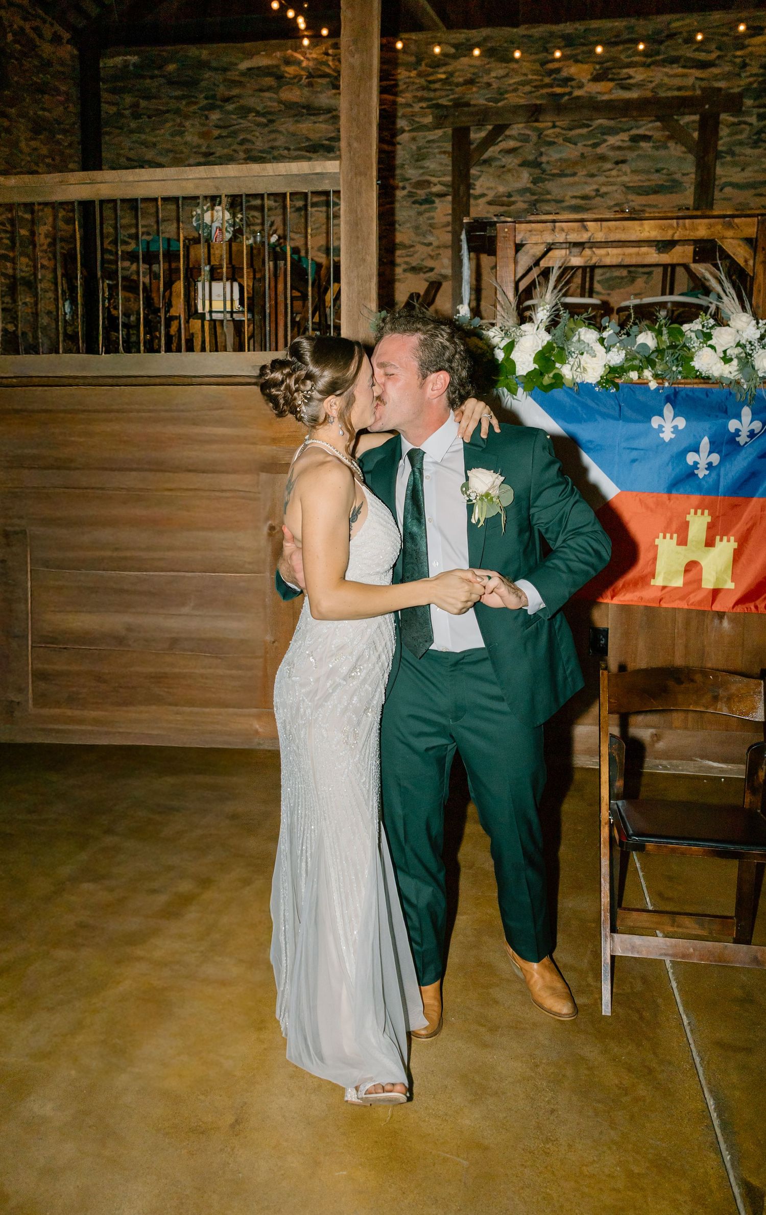 Bride and groom share a first dance kiss in a rustic barn wedding venue with string lights and floral arrangements.