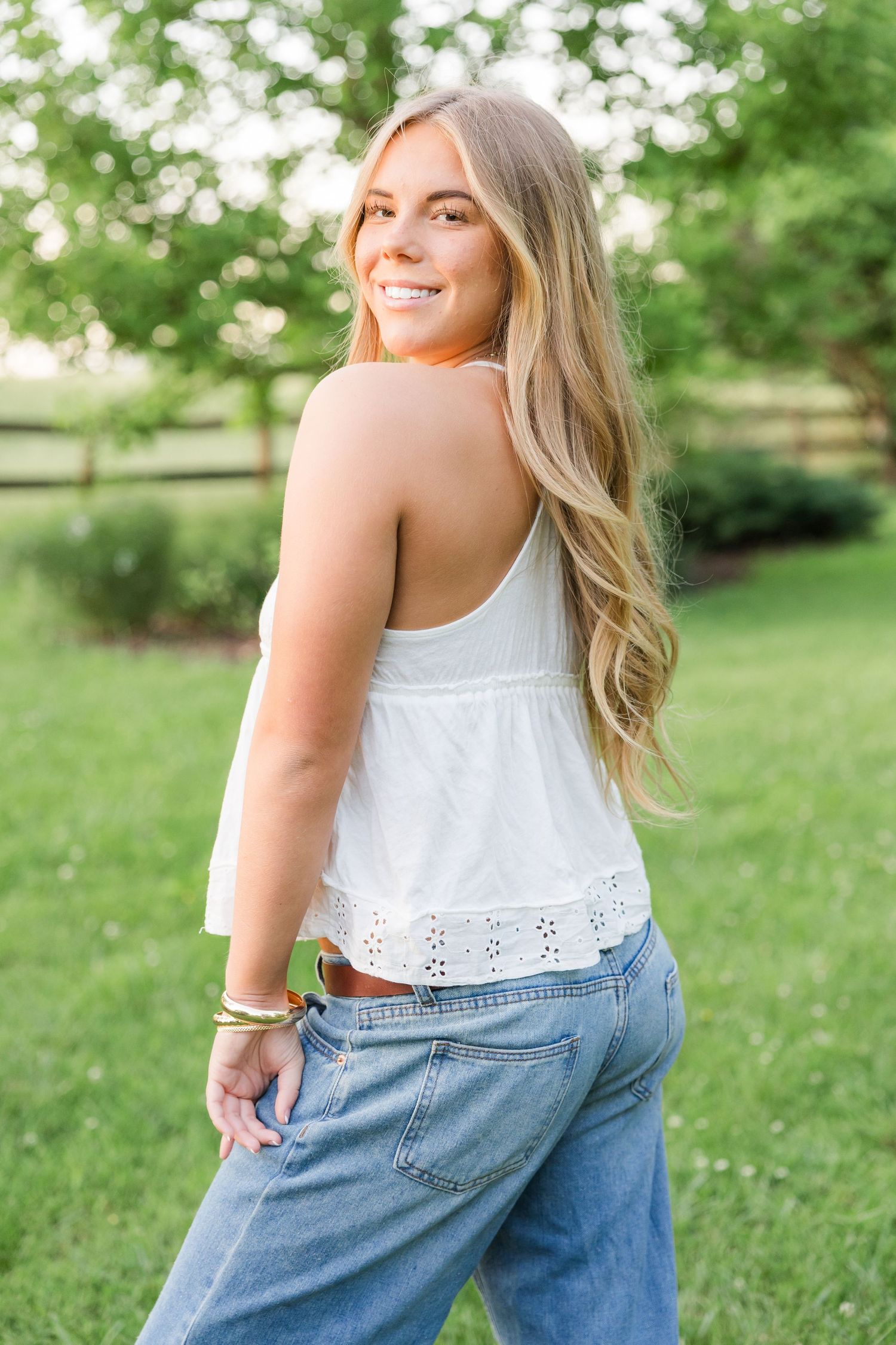 Woman in white eyelet top and jeans posing playfully in a green summer garden.