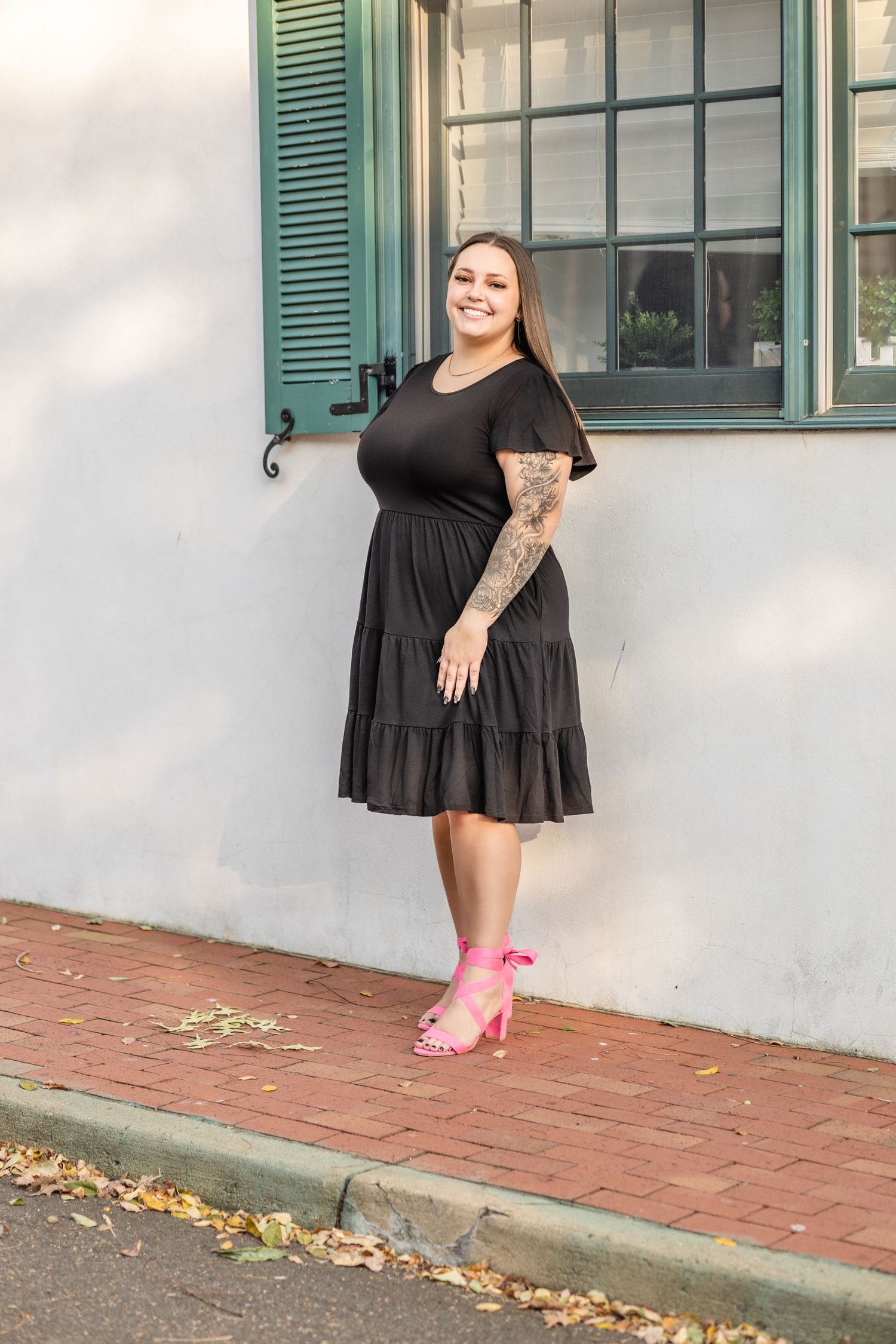 A stylish woman in a black dress and pink heels poses against a white wall with green shutters.