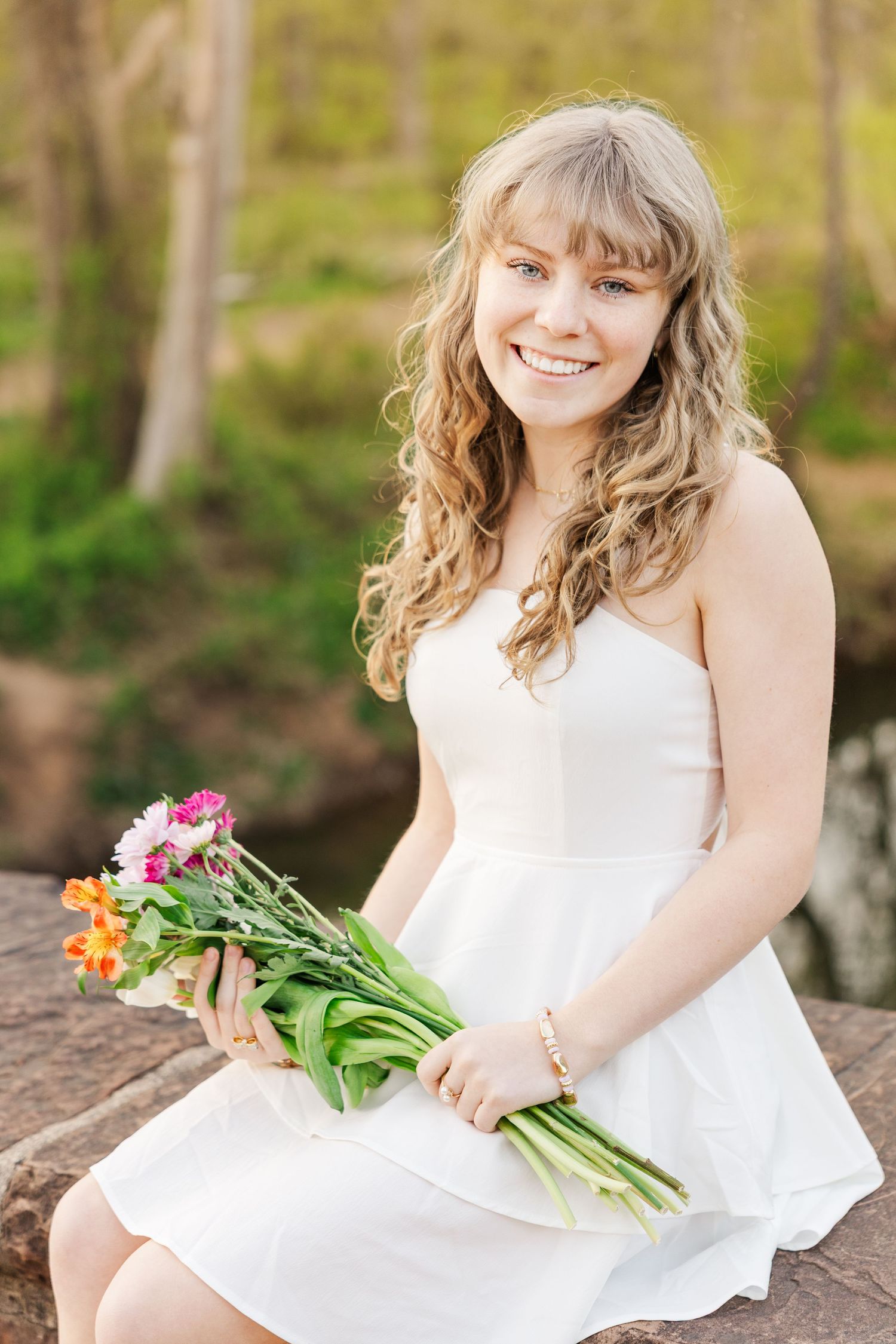A woman in a white dress holds a colorful bouquet of flowers while posing outdoors in a natural setting.