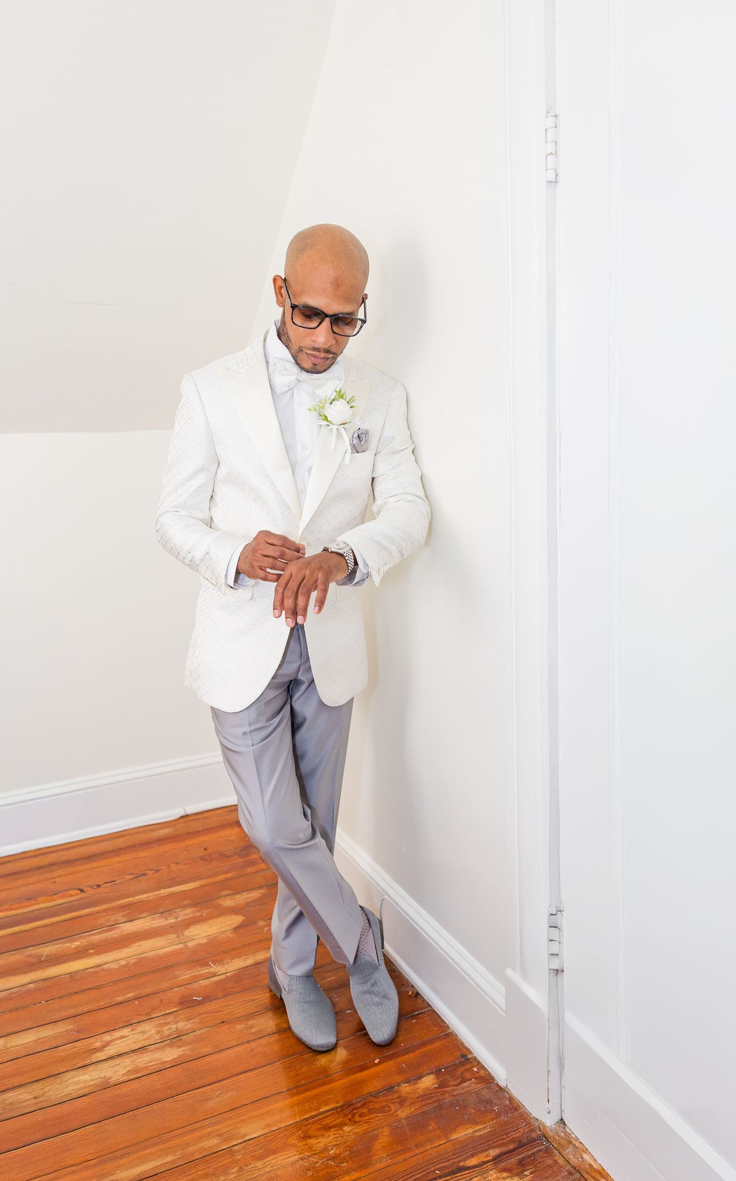 A person wearing a white blazer and gray pants stands against a white wall on hardwood floors looking downward.