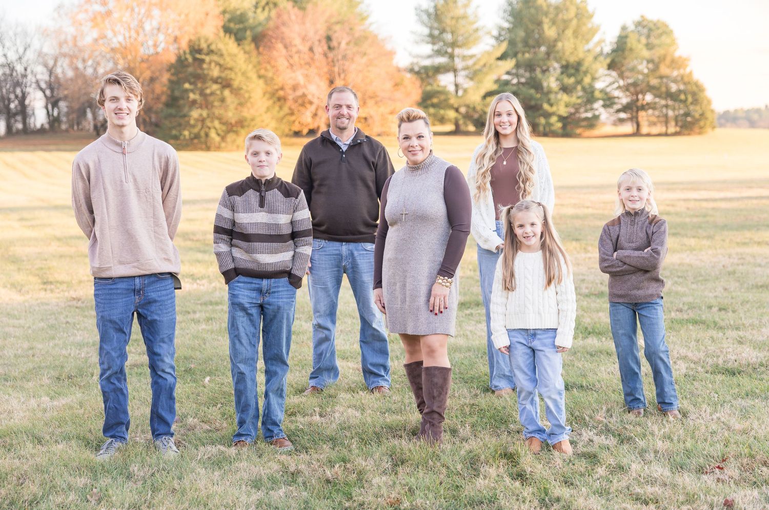 A family group stands together in a golden autumn field at sunset wearing coordinated casual outfits.