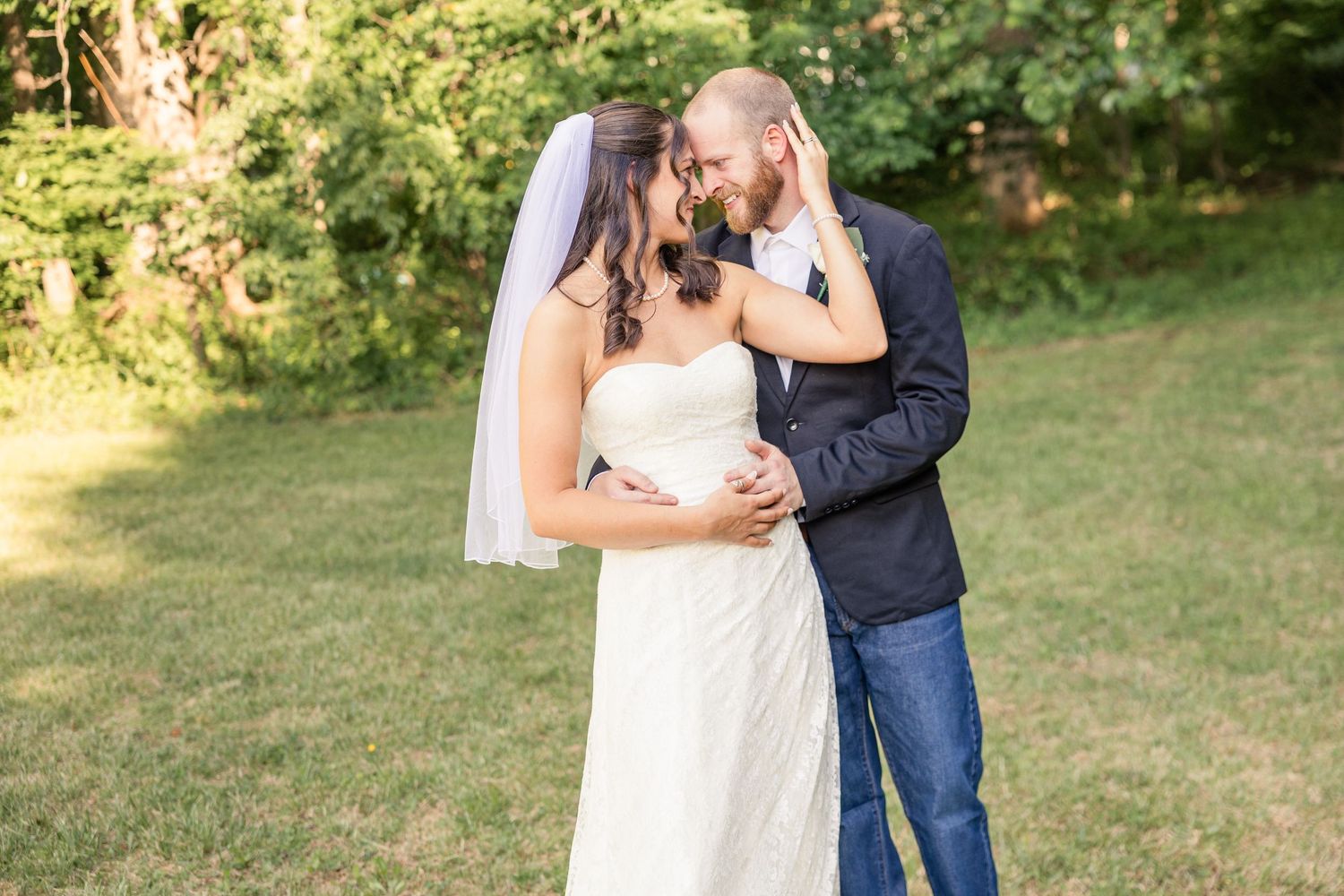 Bride in white strapless gown and veil embracing groom in navy blazer and jeans outdoors on green lawn.