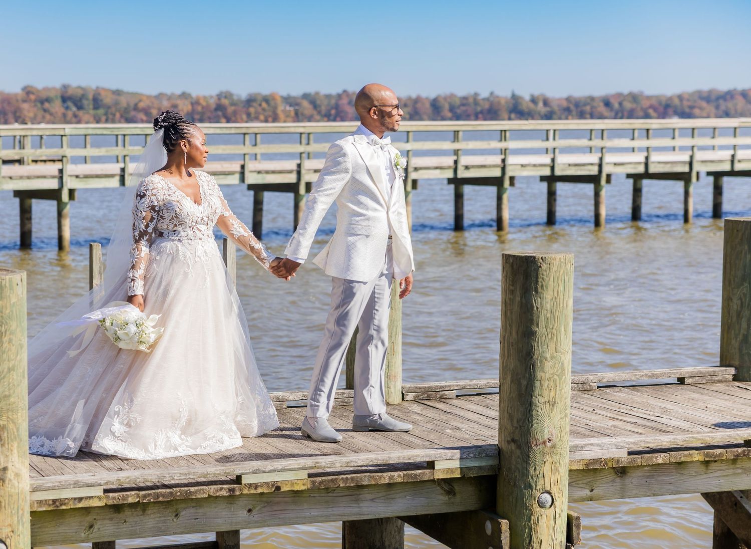 A couple in white attire poses on a wooden dock overlooking a serene lake with autumn trees in the background.