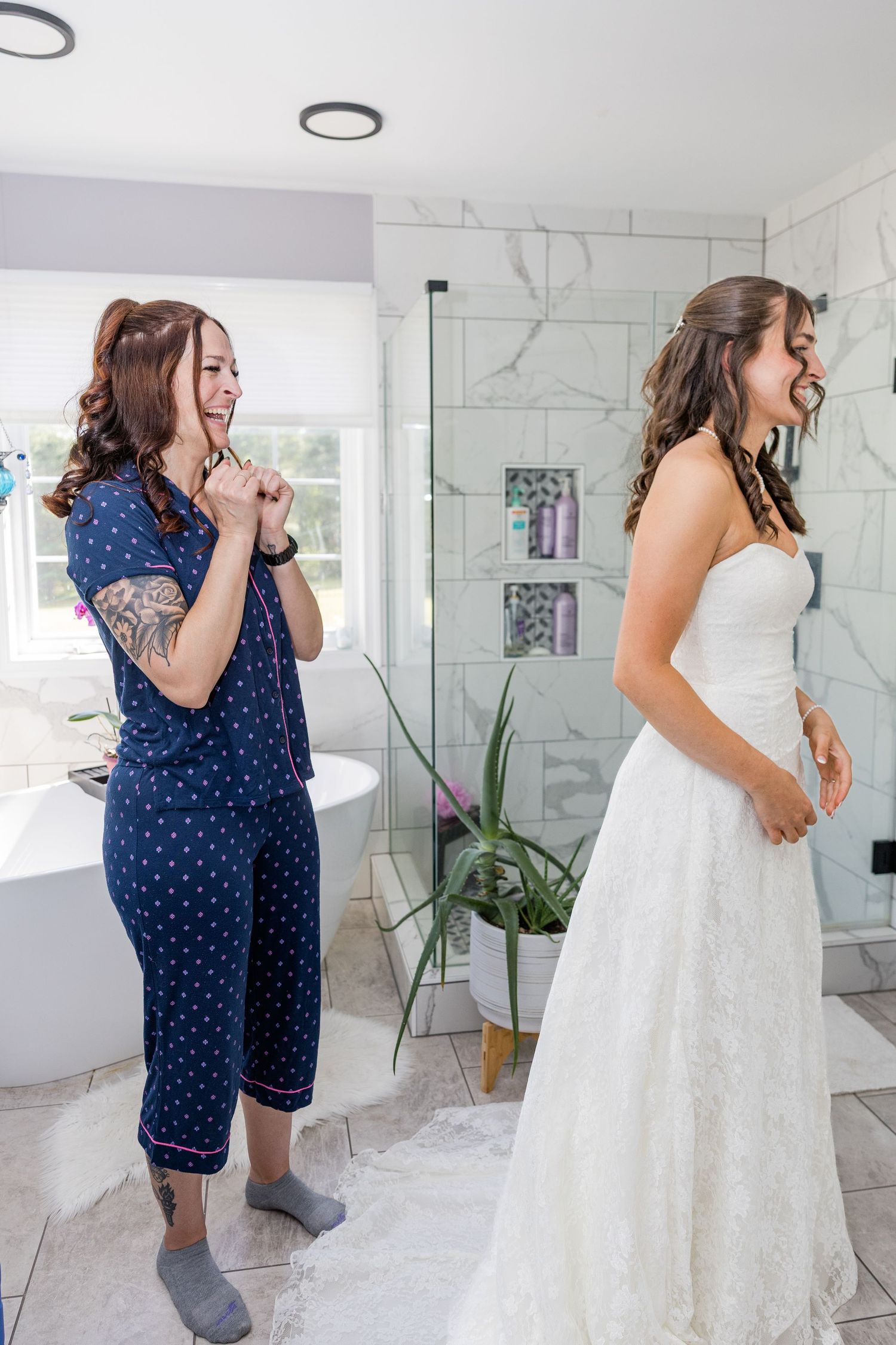 Bride in white strapless gown laughs with friend in blue pajamas during wedding morning preparations in elegant marble bathroom.