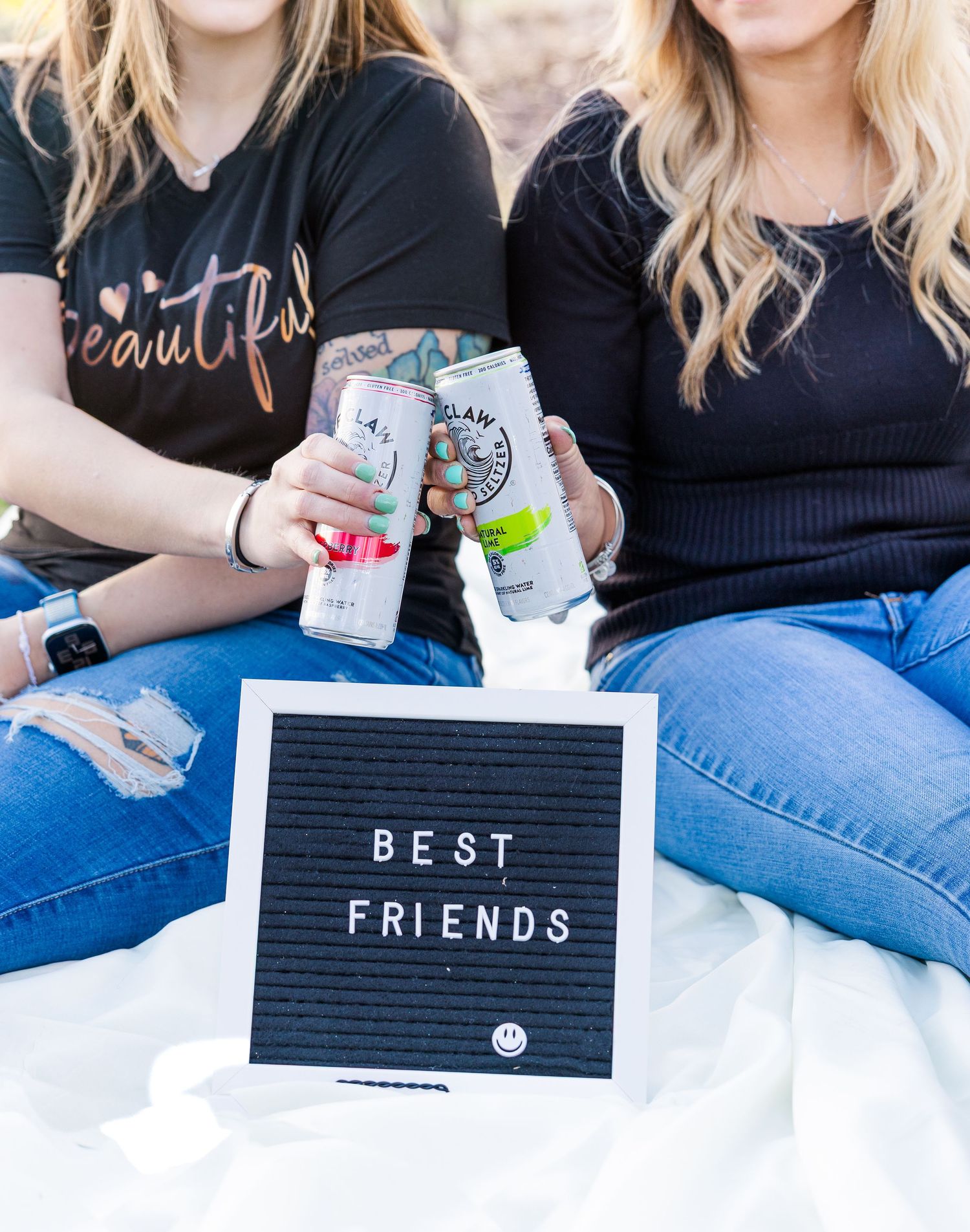 Two friends toasting with canned drinks next to a letter board that says Best Friends.