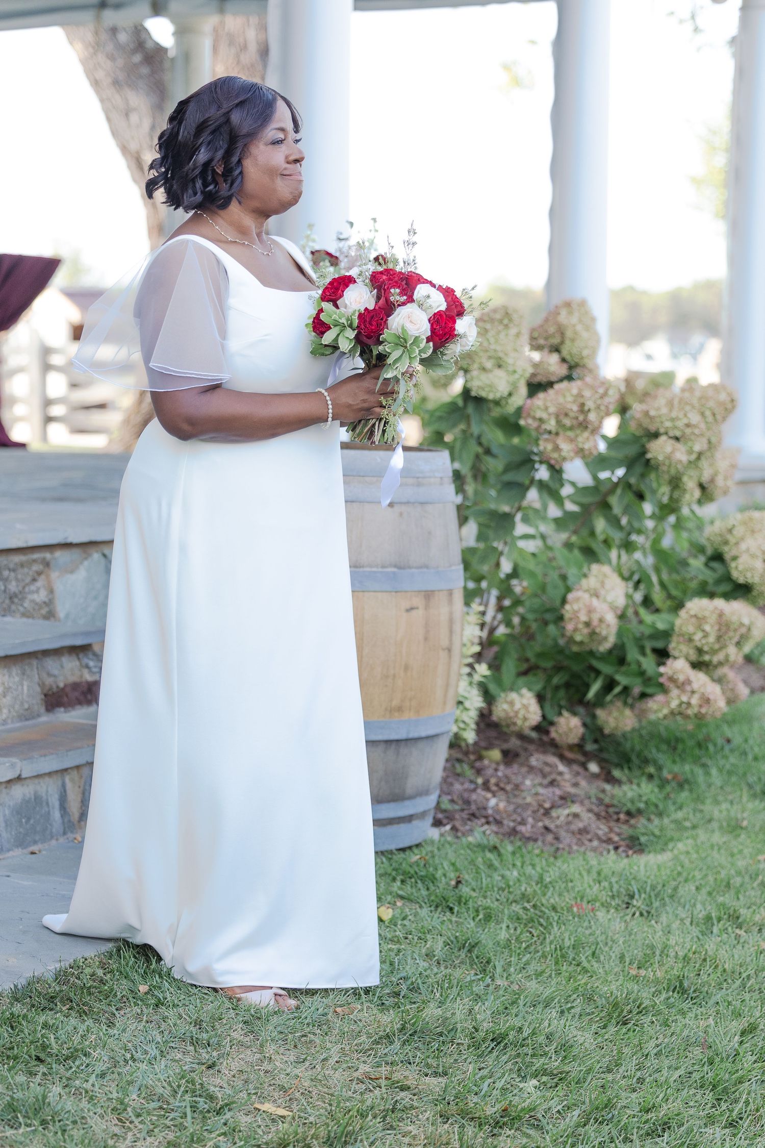 A bride in a white dress holds a red and white rose bouquet against a garden backdrop.