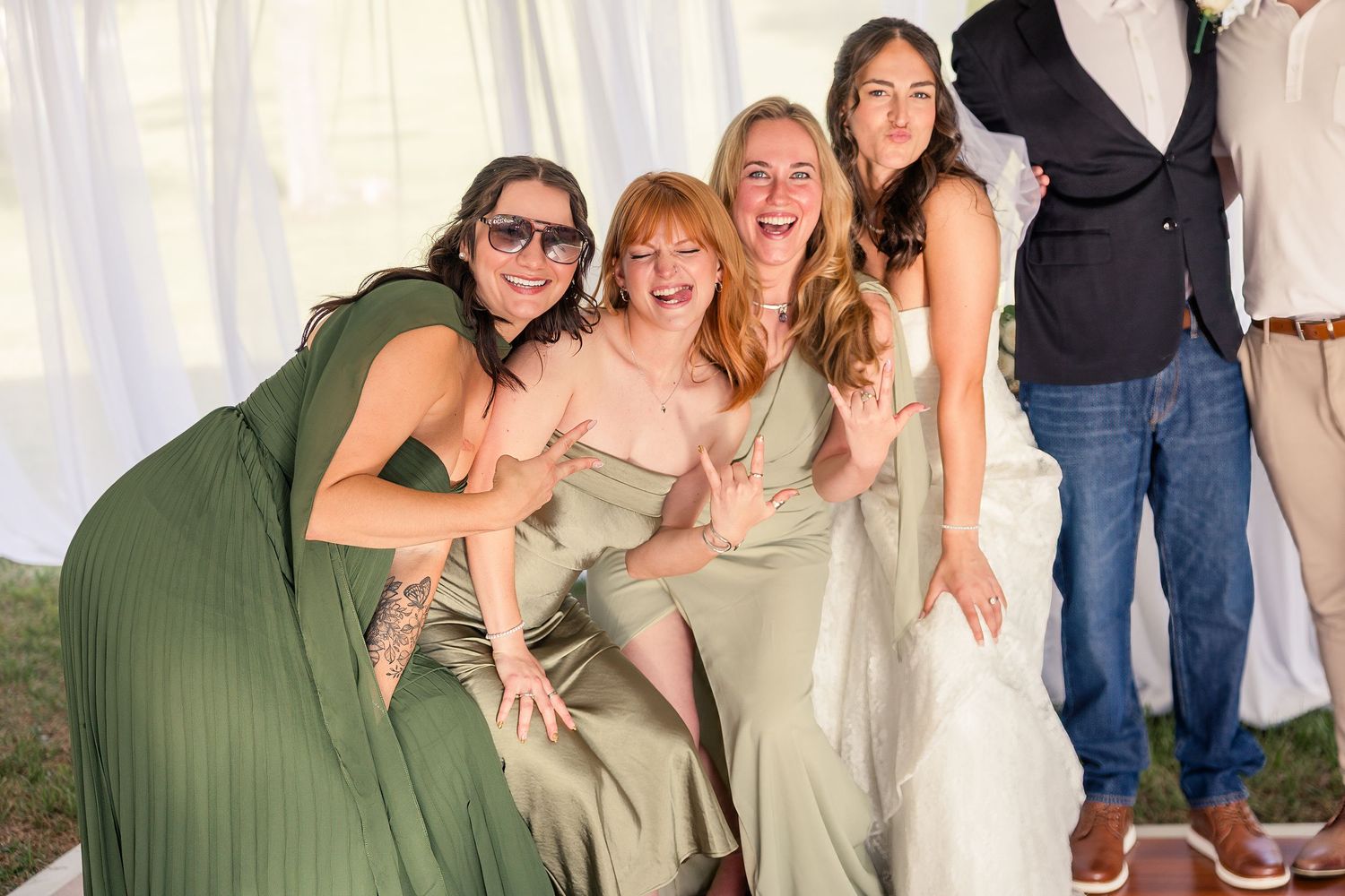 Four women in sage green dresses laugh and pose together at an outdoor wedding reception.