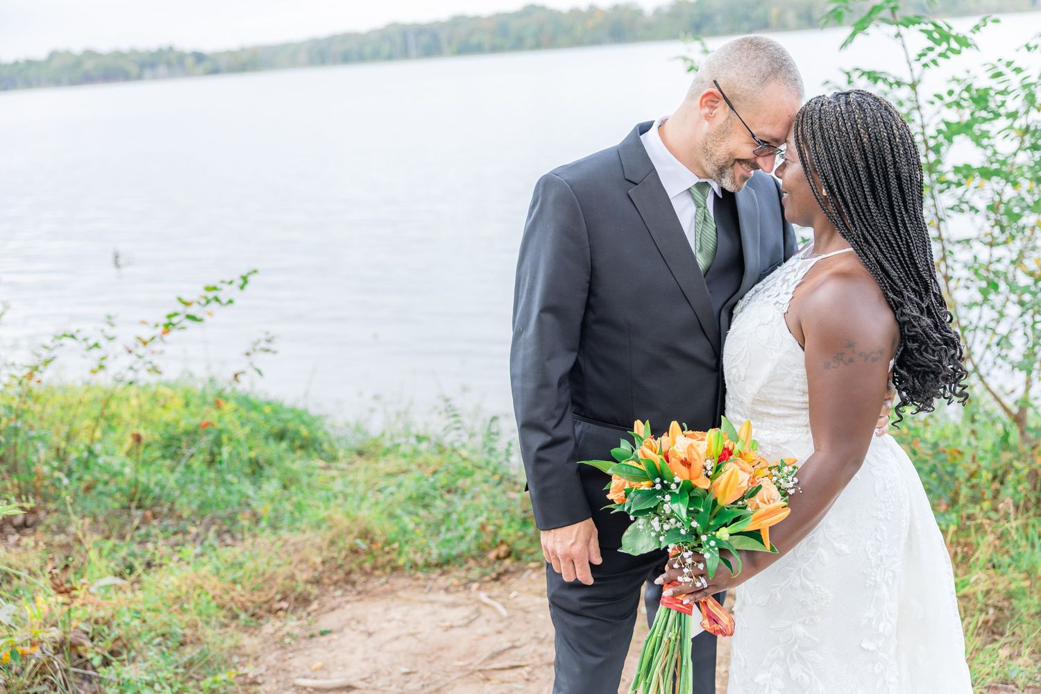 Wedding couple poses near lakeside with orange floral bouquet.