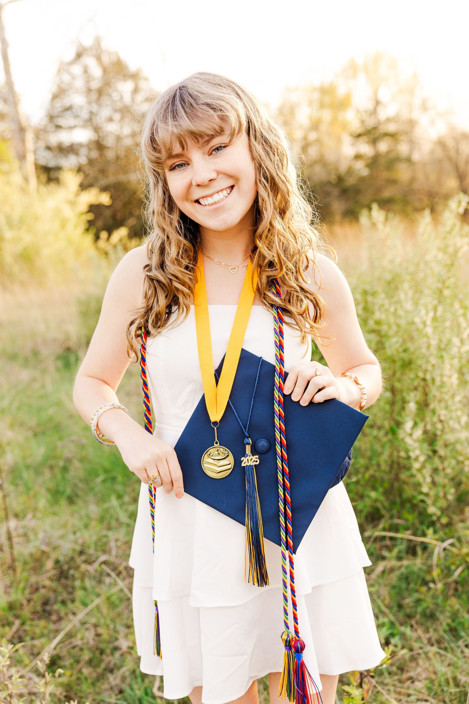 Graduate wearing white dress and blue stole with gold honors cords in sunny field.