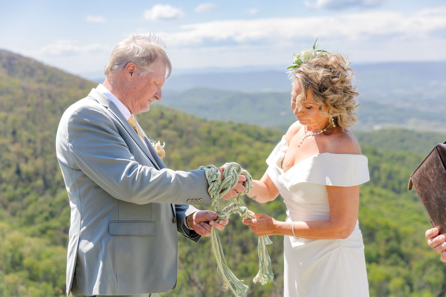 Bride and groom performing handfasting ceremony outdoors with mountain views in background.