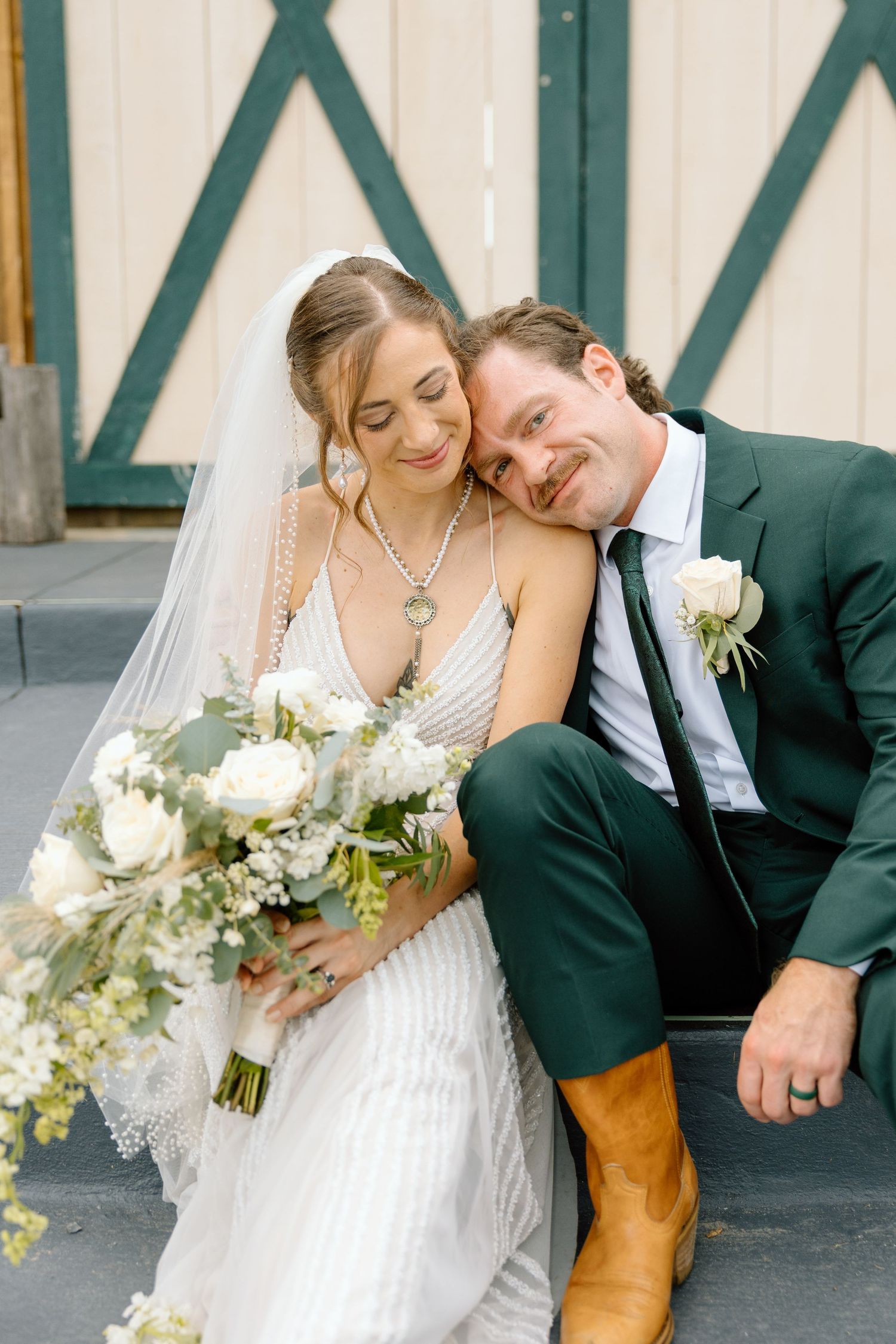 Bride in white gown with veil holds bouquet as groom in green suit rests head on hers on wedding day.