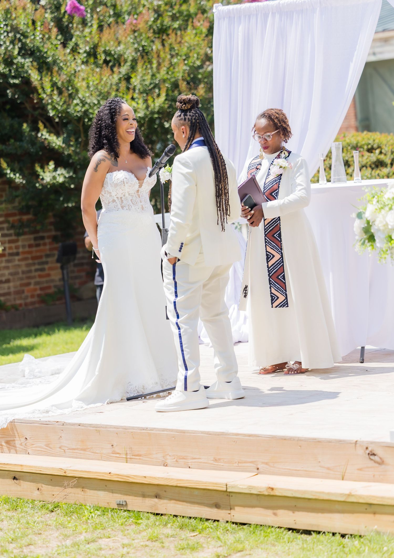 An outdoor wedding ceremony setup with white drapery and wooden platform against a garden backdrop.