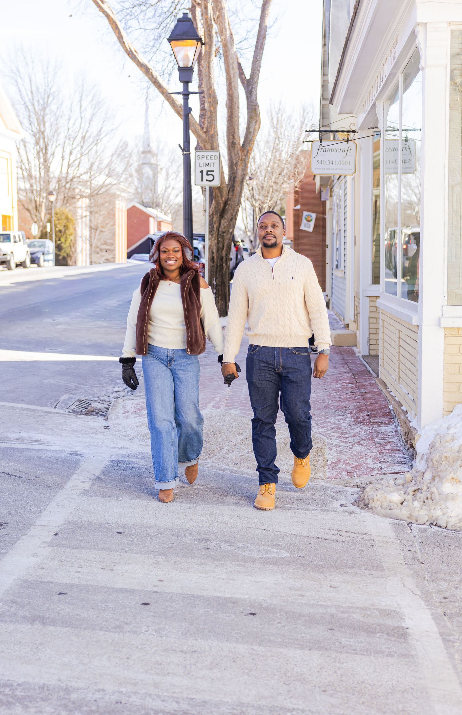 A couple walks down a snowy small town sidewalk together on a winter day.