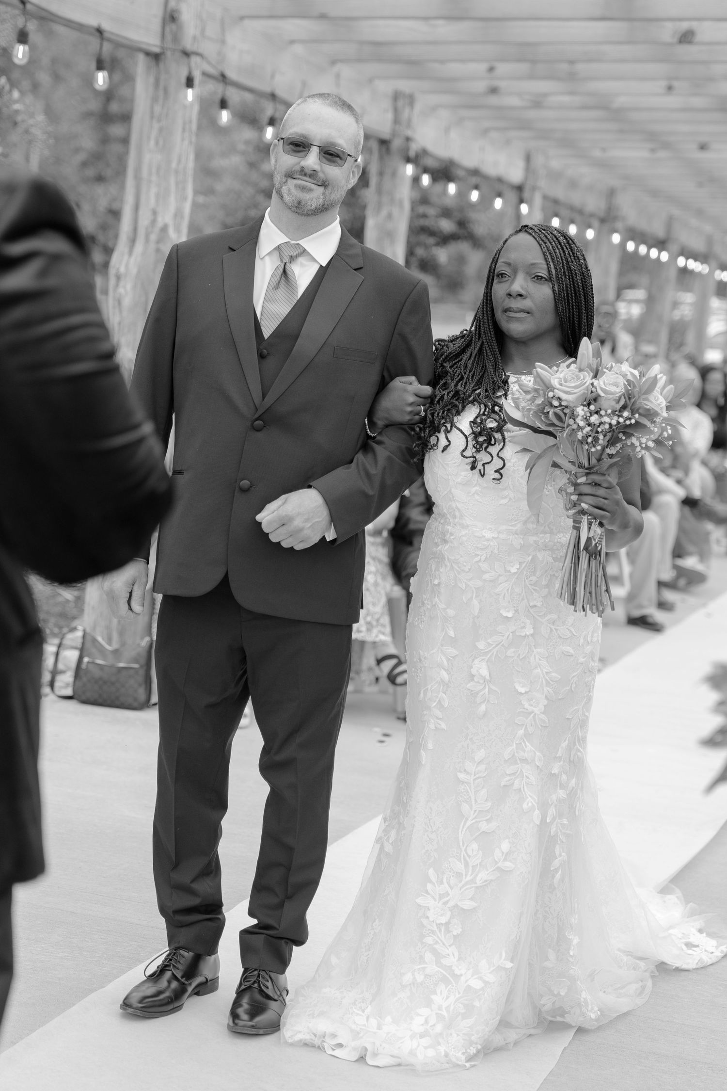 Black and white photo of a couple walking down an aisle with string lights overhead.