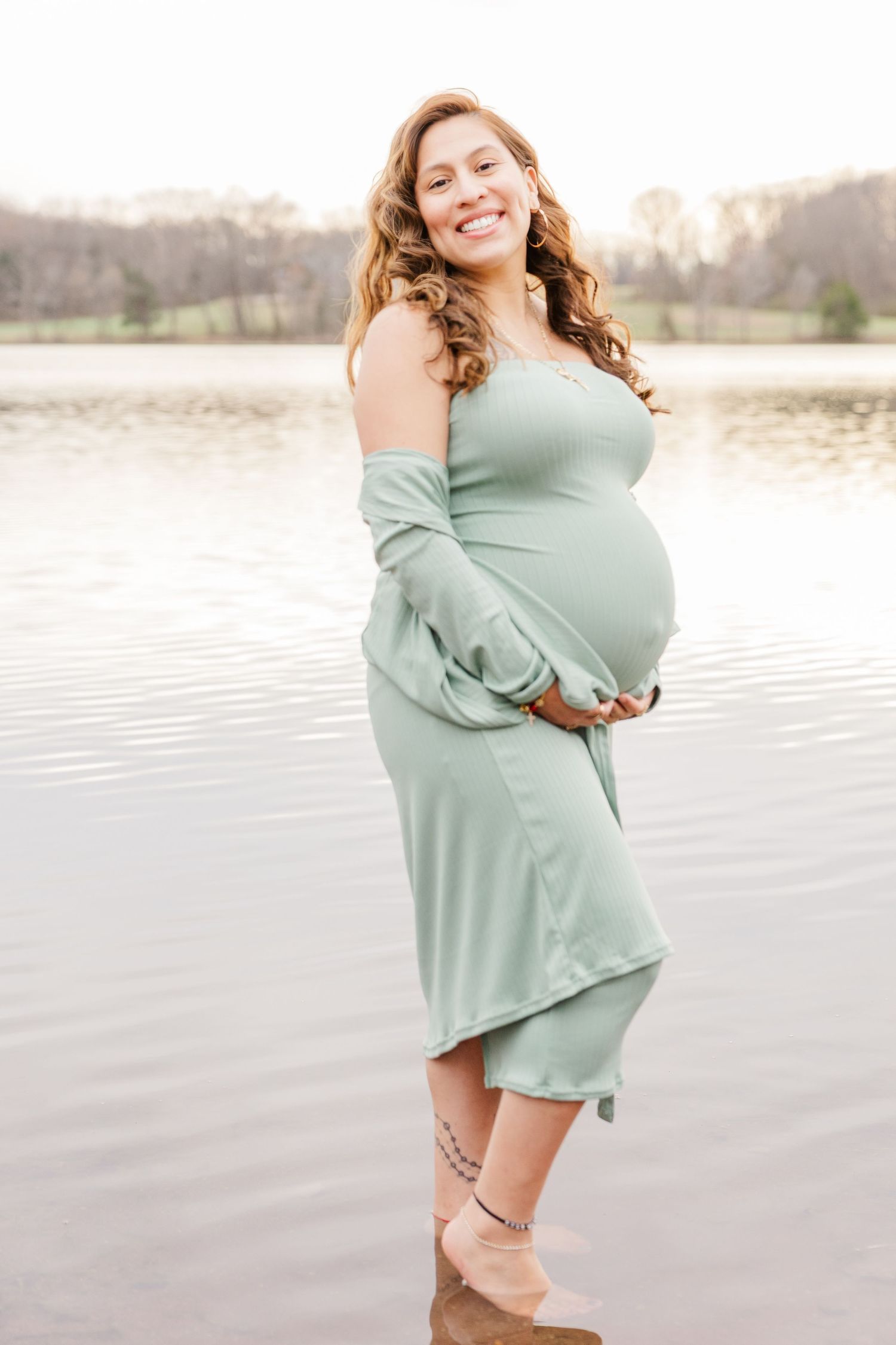 A maternity photoshoot by a lake featuring a woman in a flowing mint green dress at sunset.