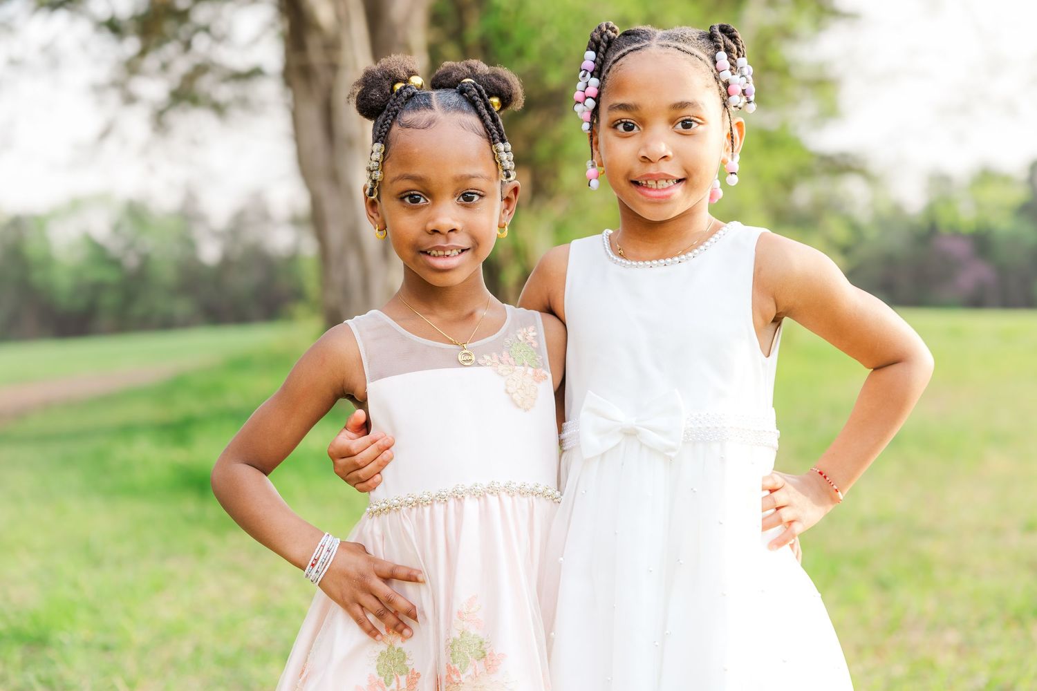 Two young girls in white and pink dresses pose together outdoors on a sunny day.