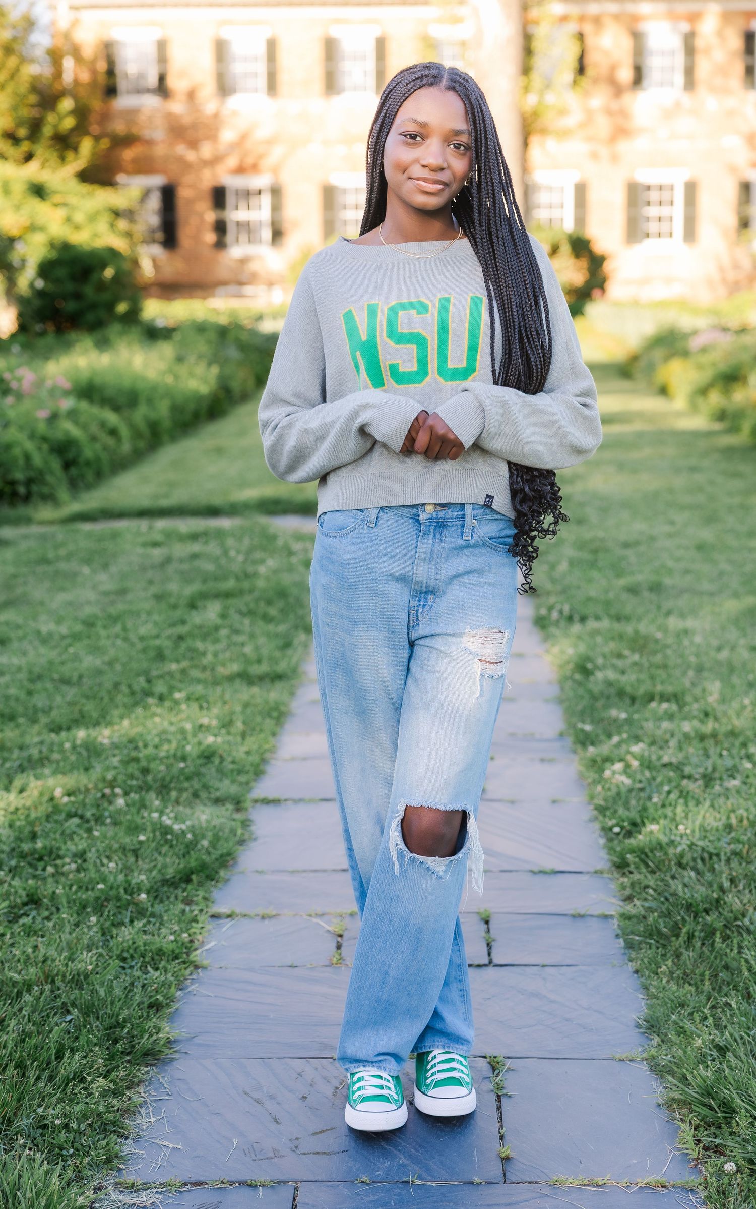 Student wearing NSU sweatshirt and ripped jeans stands on stone path with brick building in background.
