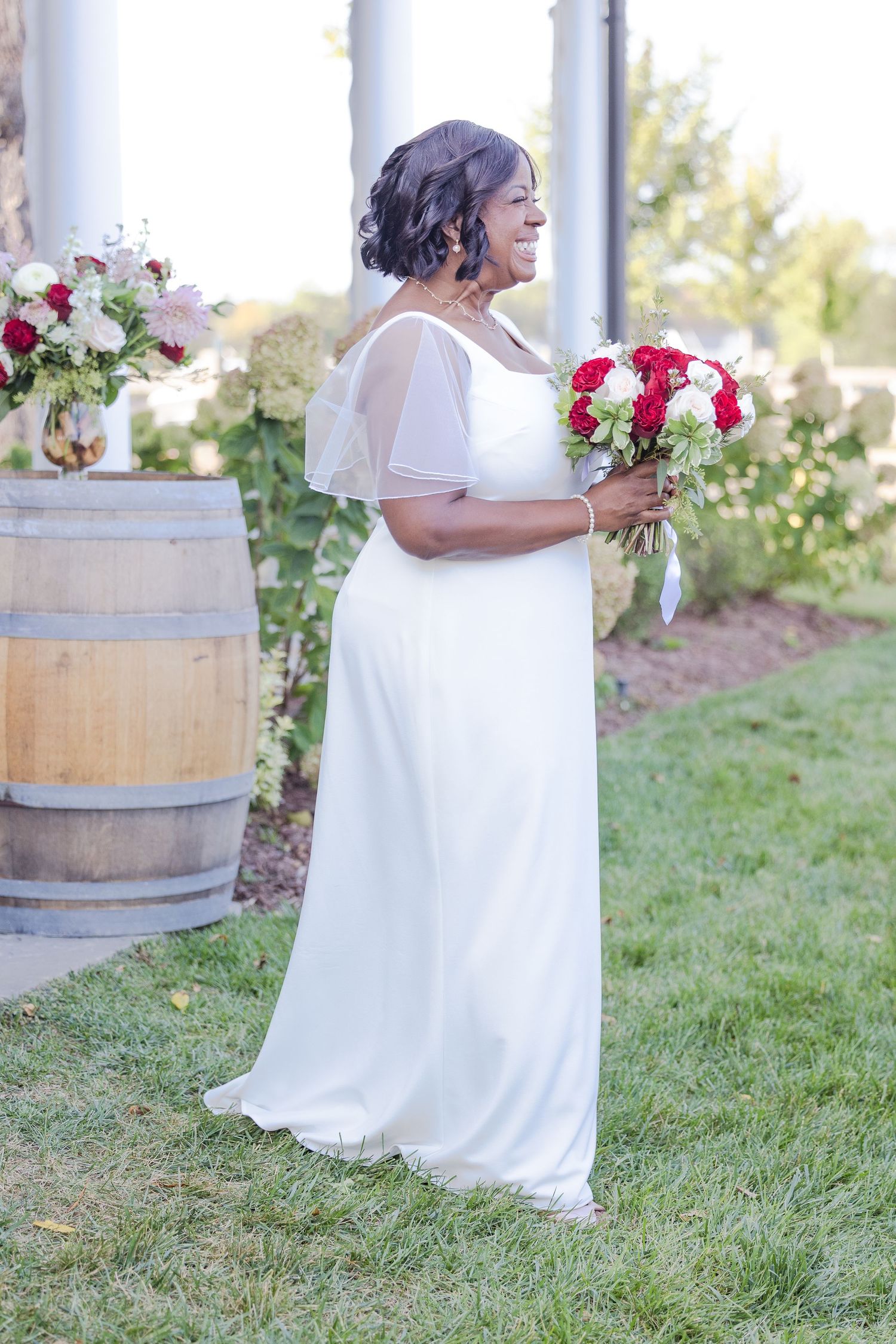 A bride in a flowing white gown poses near wooden barrels in an outdoor garden setting.