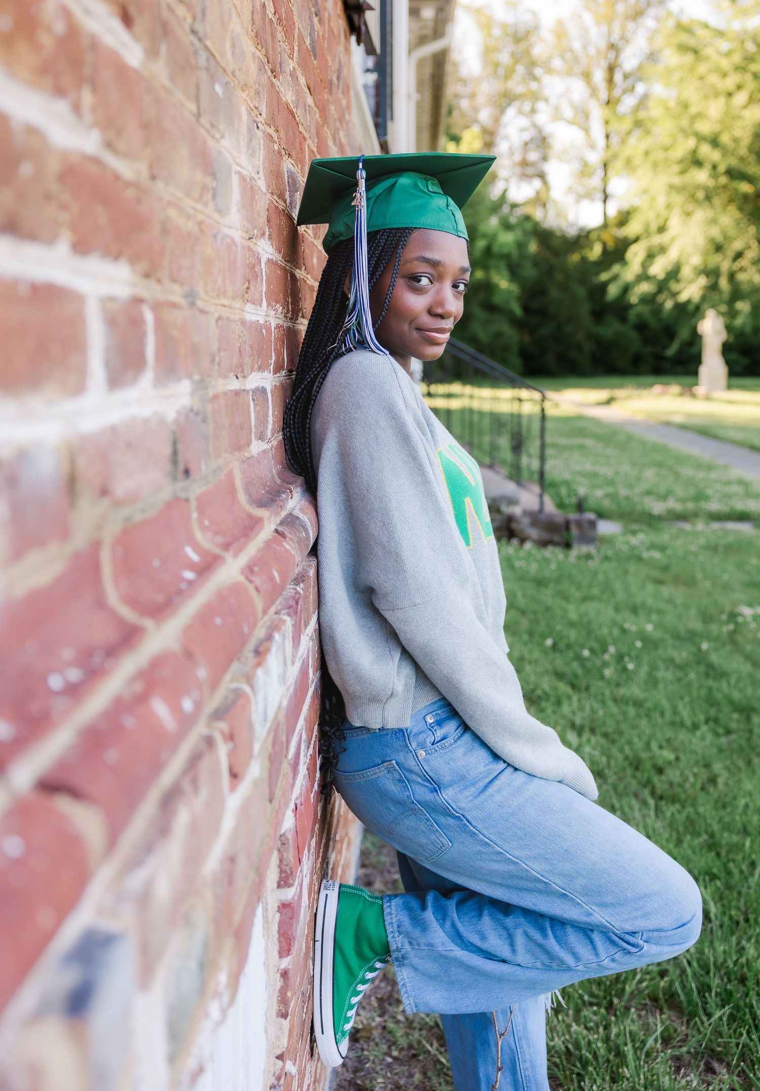 Graduate in green cap and gown leans against brick wall outdoors.