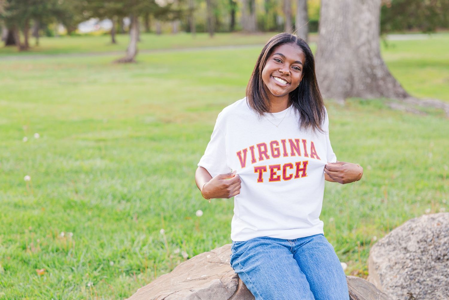 A person in Virginia Tech apparel poses casually on grass near trees in a park setting.