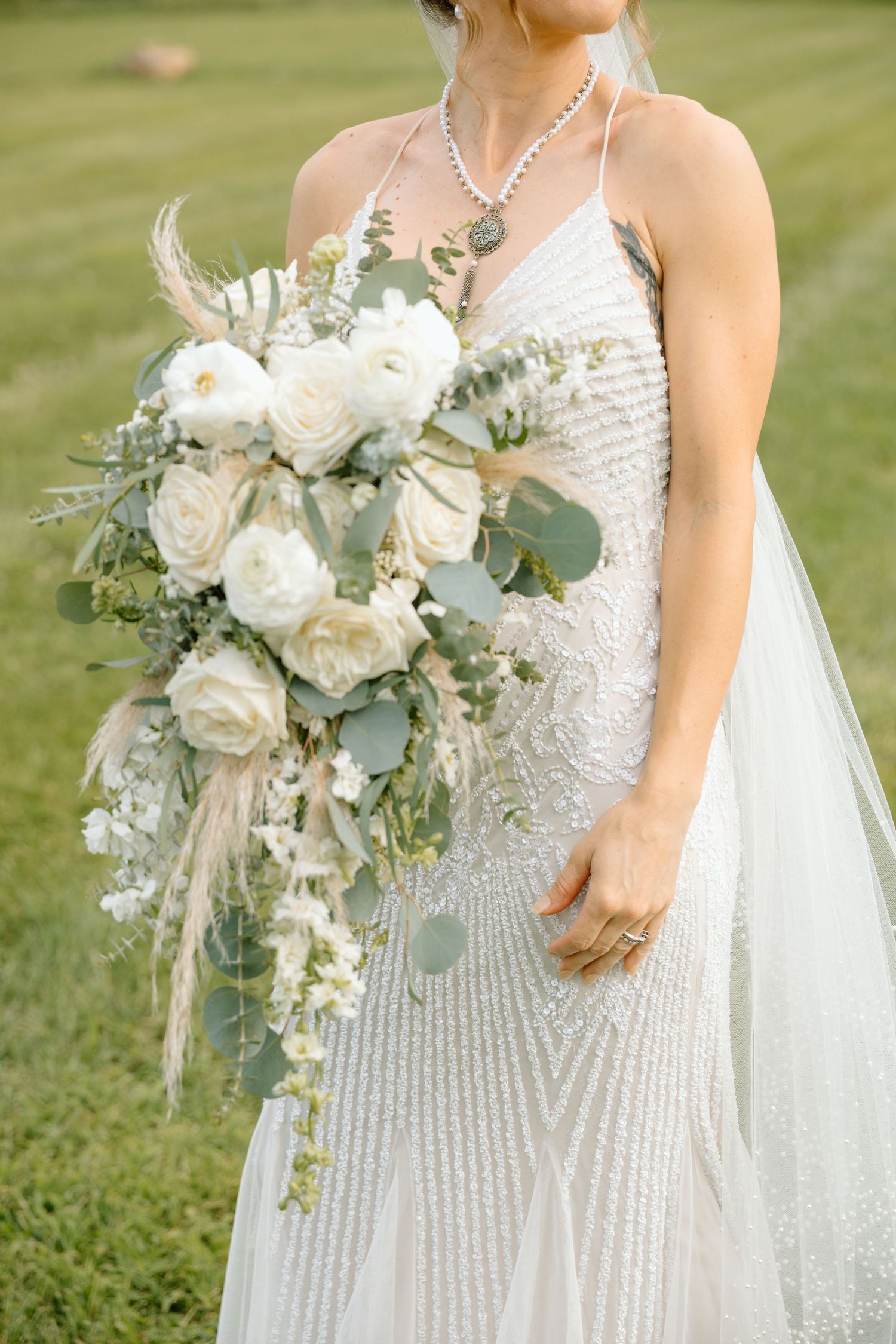 Bride in beaded white gown holding cascading bouquet of white roses and eucalyptus with pampas grass accents.