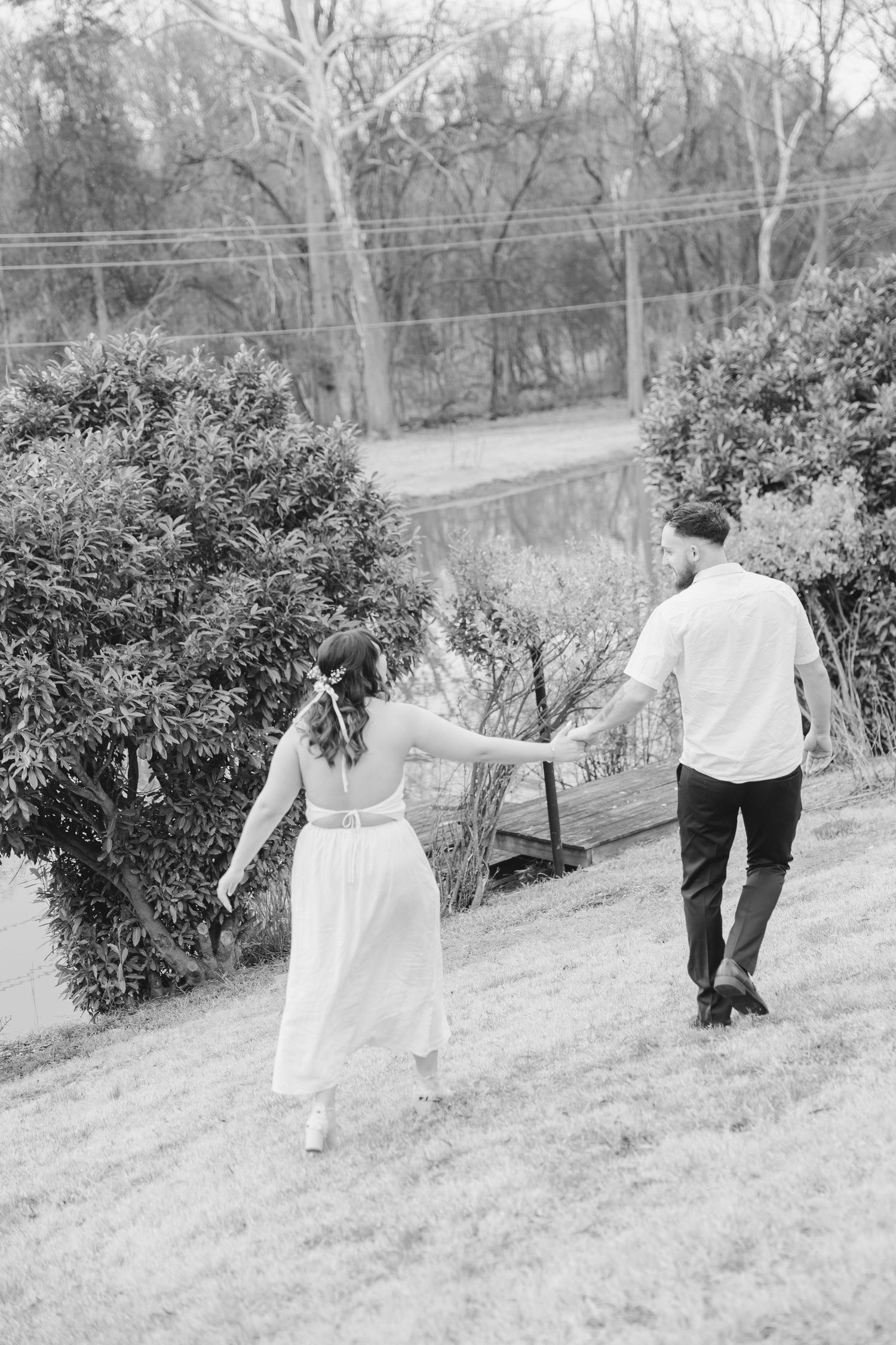 A couple in formal attire walks hand in hand up a grassy hill surrounded by trees in a black and white photograph.