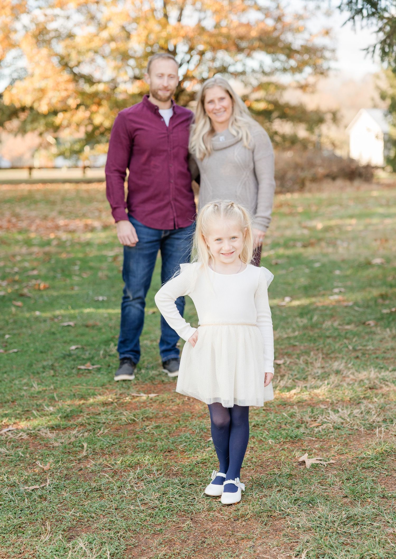 A young child in a white dress walks across an autumn lawn with family members in burgundy and gray clothing.