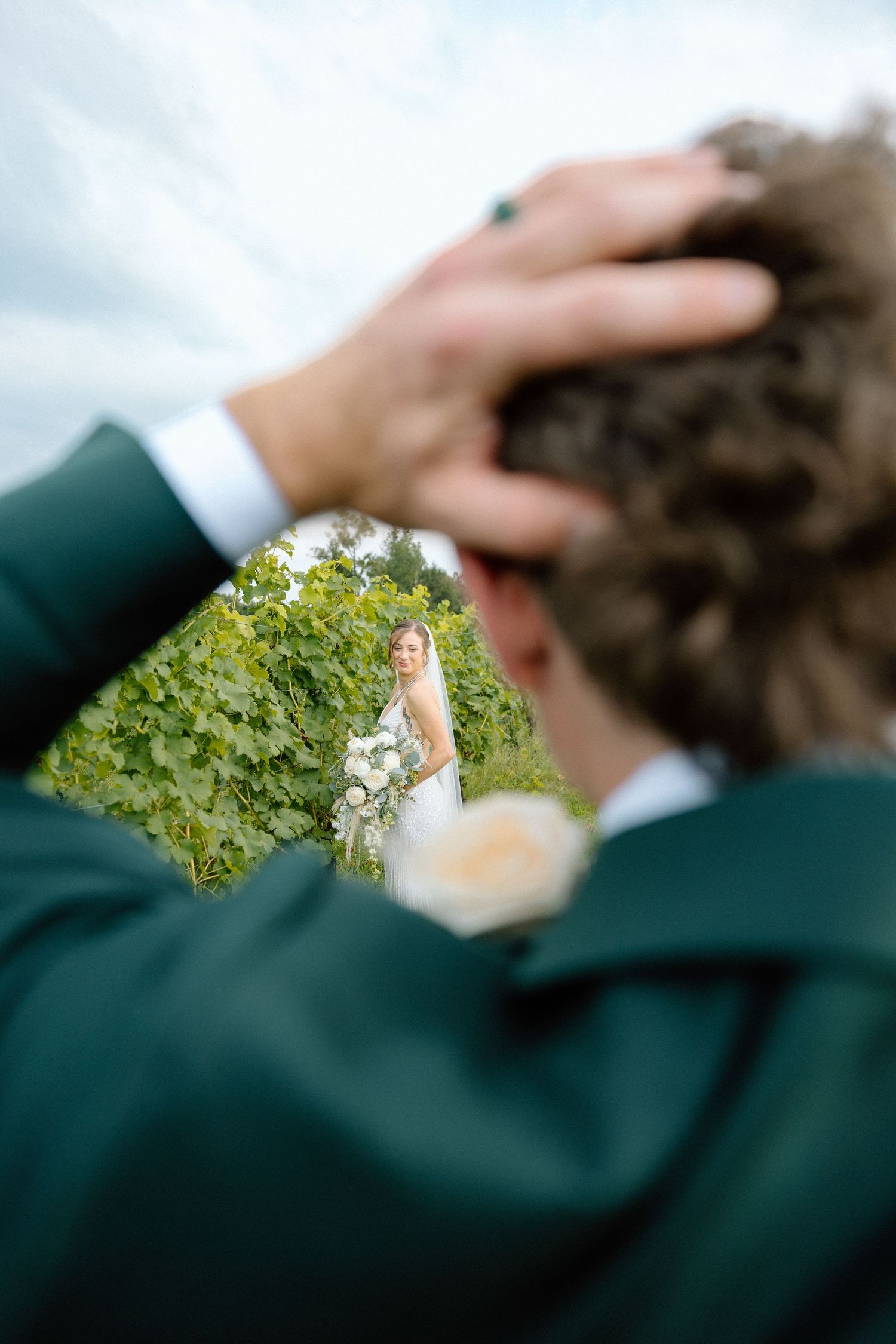 Groom in green suit touching his curly hair while bride in white dress holds bouquet among lush greenery.