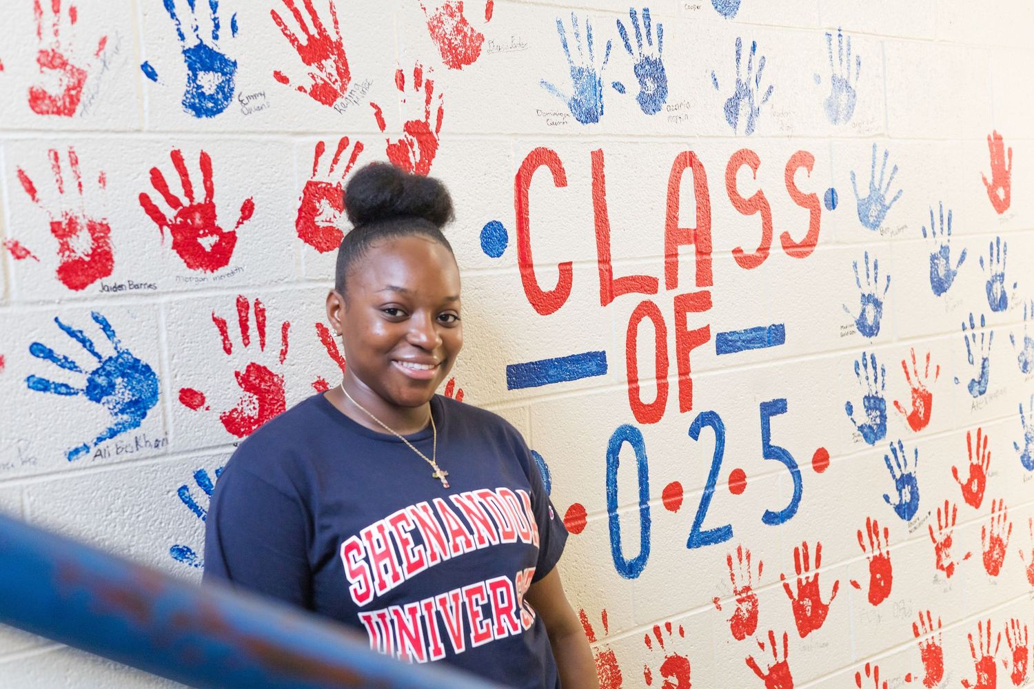 A person poses against a wall decorated with colorful handprints and graduation year text.
