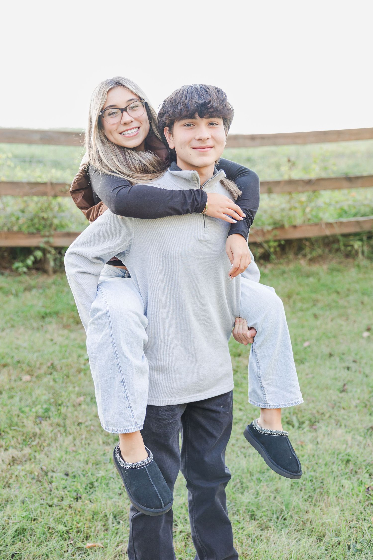A playful couple in casual attire embraces while standing in a grassy field on a bright day.