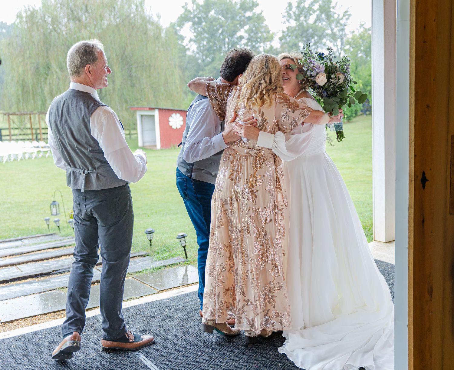 Wedding party poses outside a rustic venue with white floral arch and natural greenery backdrop.