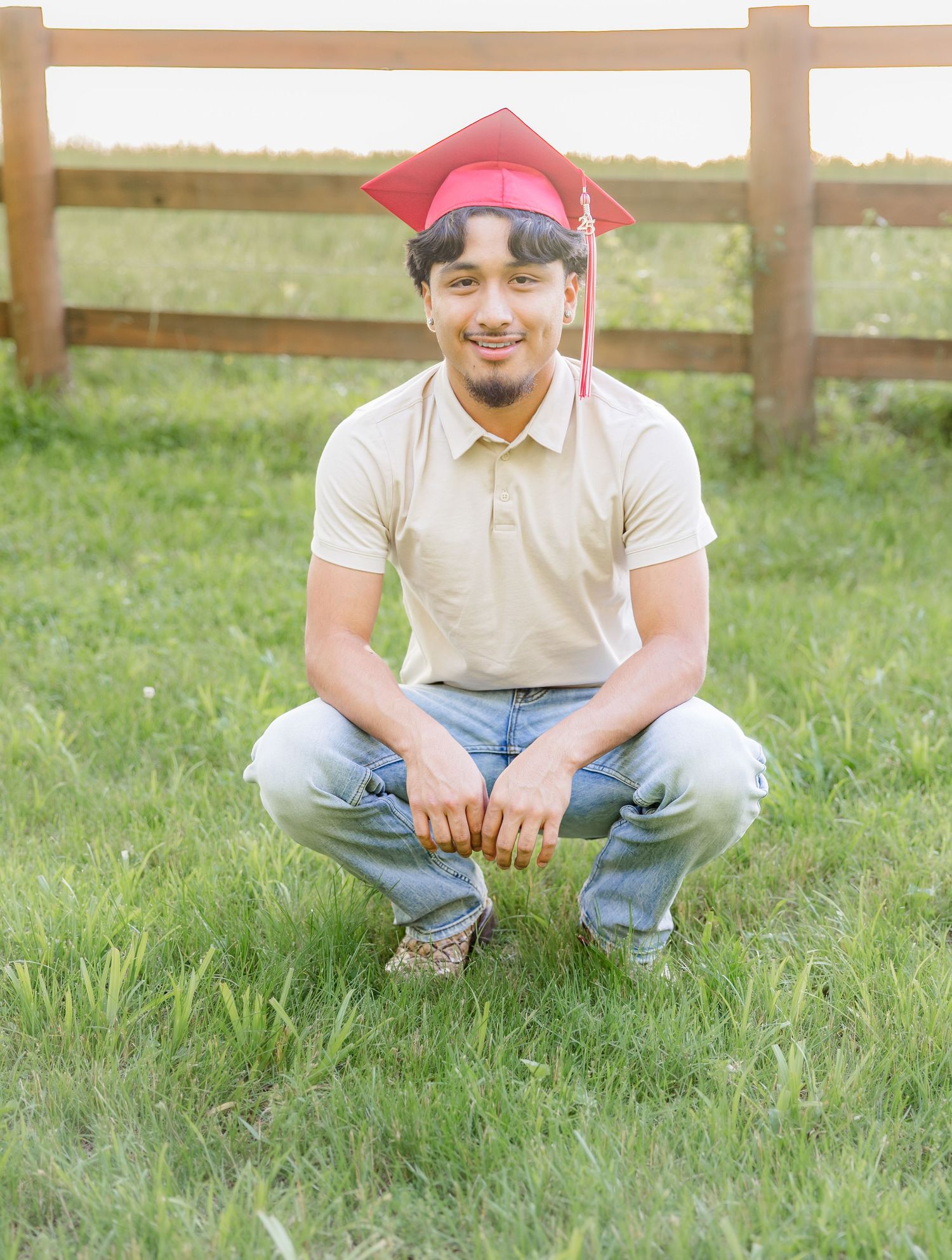 Person wearing red cap and casual attire sitting in grassy field with wooden fence background.