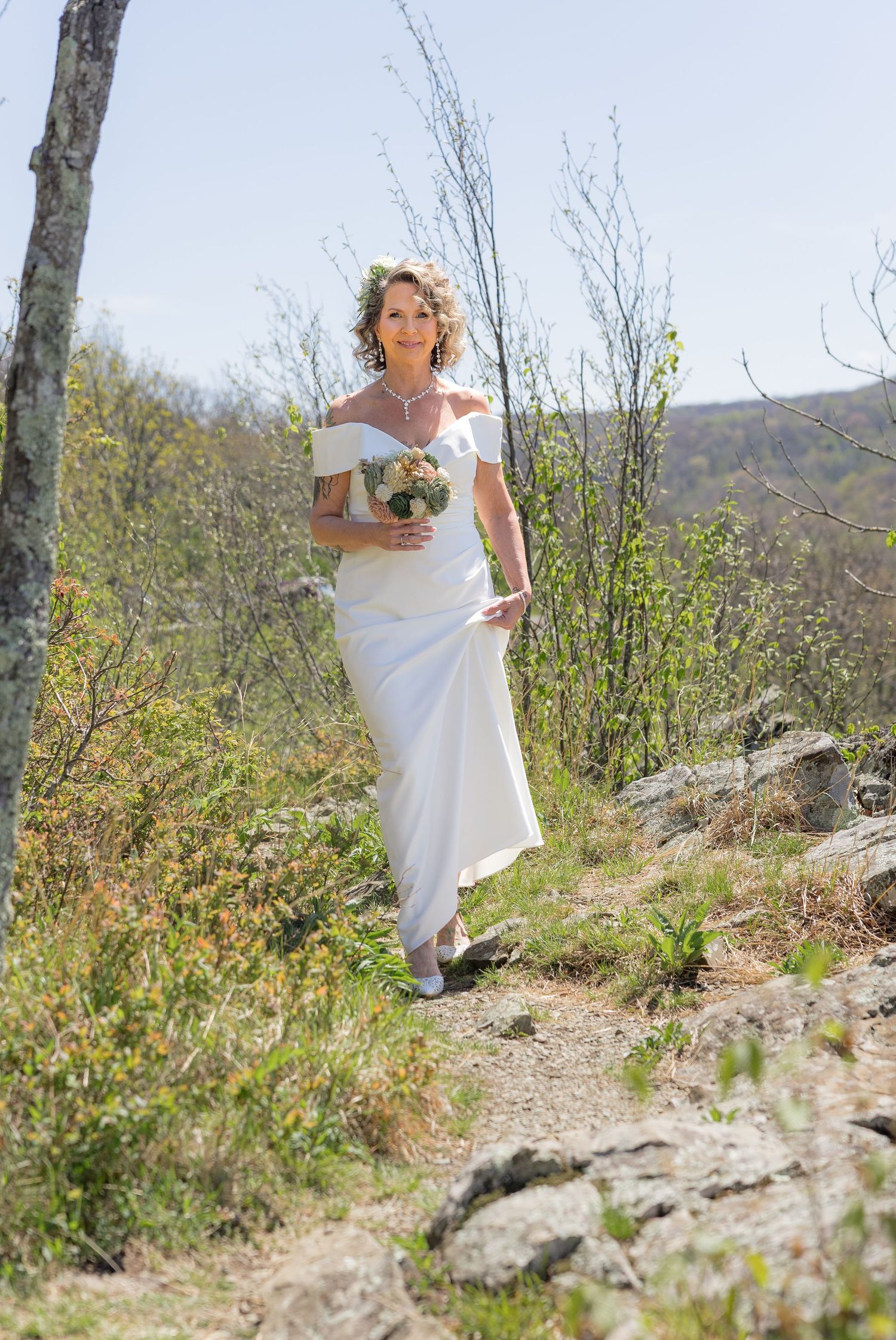 Bride in white off-shoulder gown holding bouquet walks rocky outdoor path surrounded by spring trees and hills.