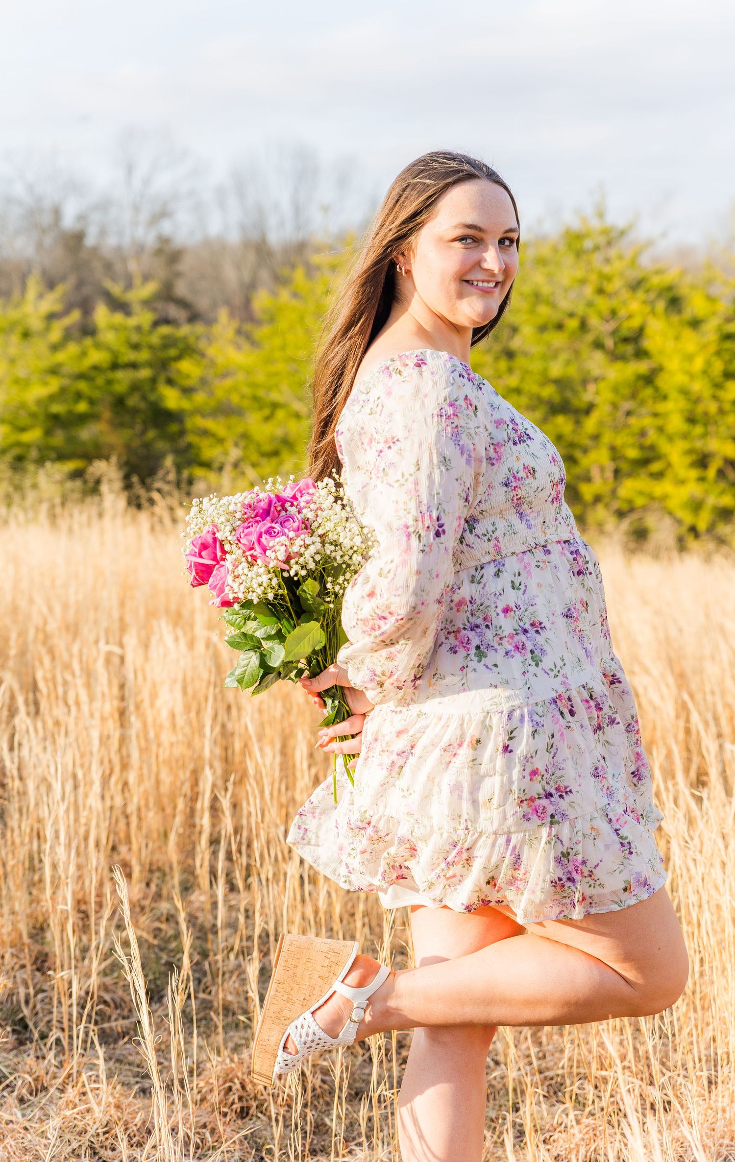 Woman in floral dress dances through golden field holding pink flowers at sunset.
