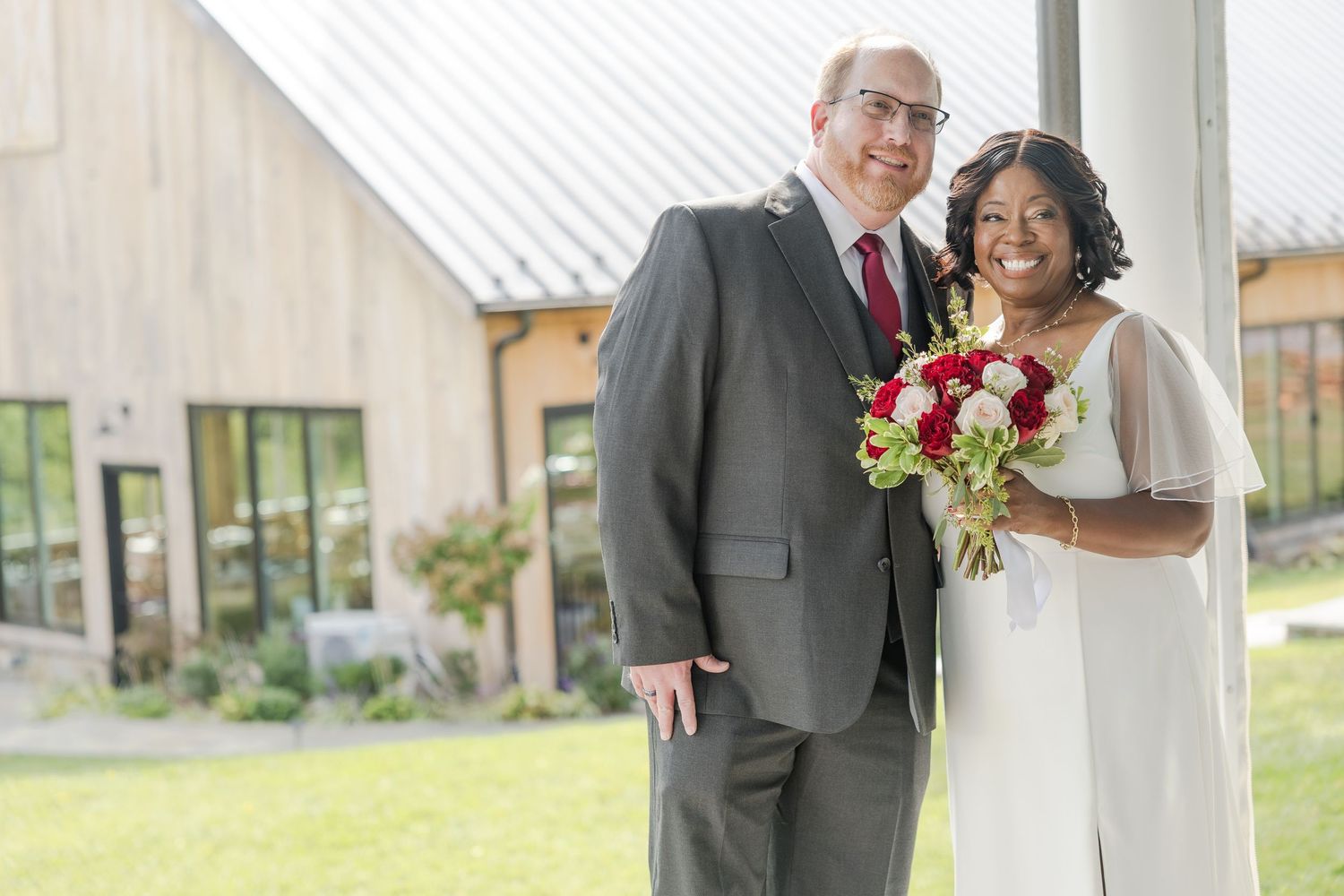 Wedding couple poses outdoors in formal attire with red and white bouquet.