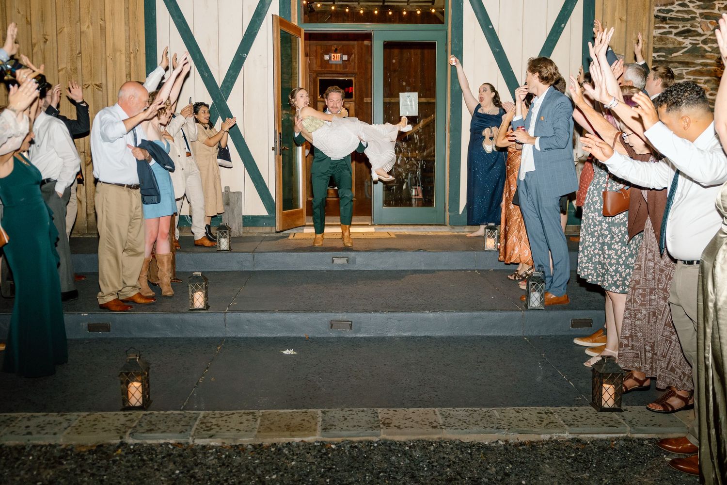 Newlywed groom carries bride down steps as wedding guests cheer and raise hands outside rustic barn venue.