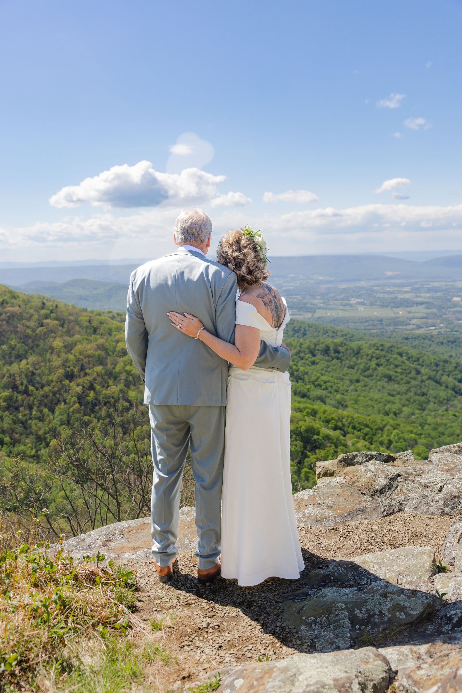 Wedding couple embraces while gazing at stunning green mountain valley landscape from rocky overlook.