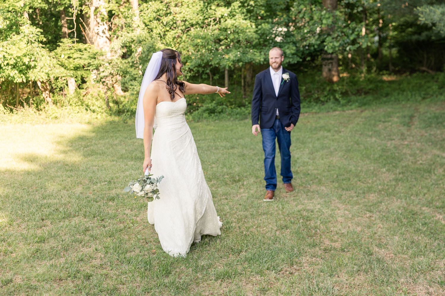 Bride in white gown points at groom in navy blazer and jeans during outdoor first look on green lawn.