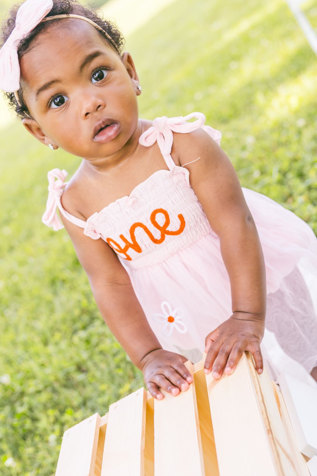 Smiling baby in pink and orange dress poses outdoors on sunny day.