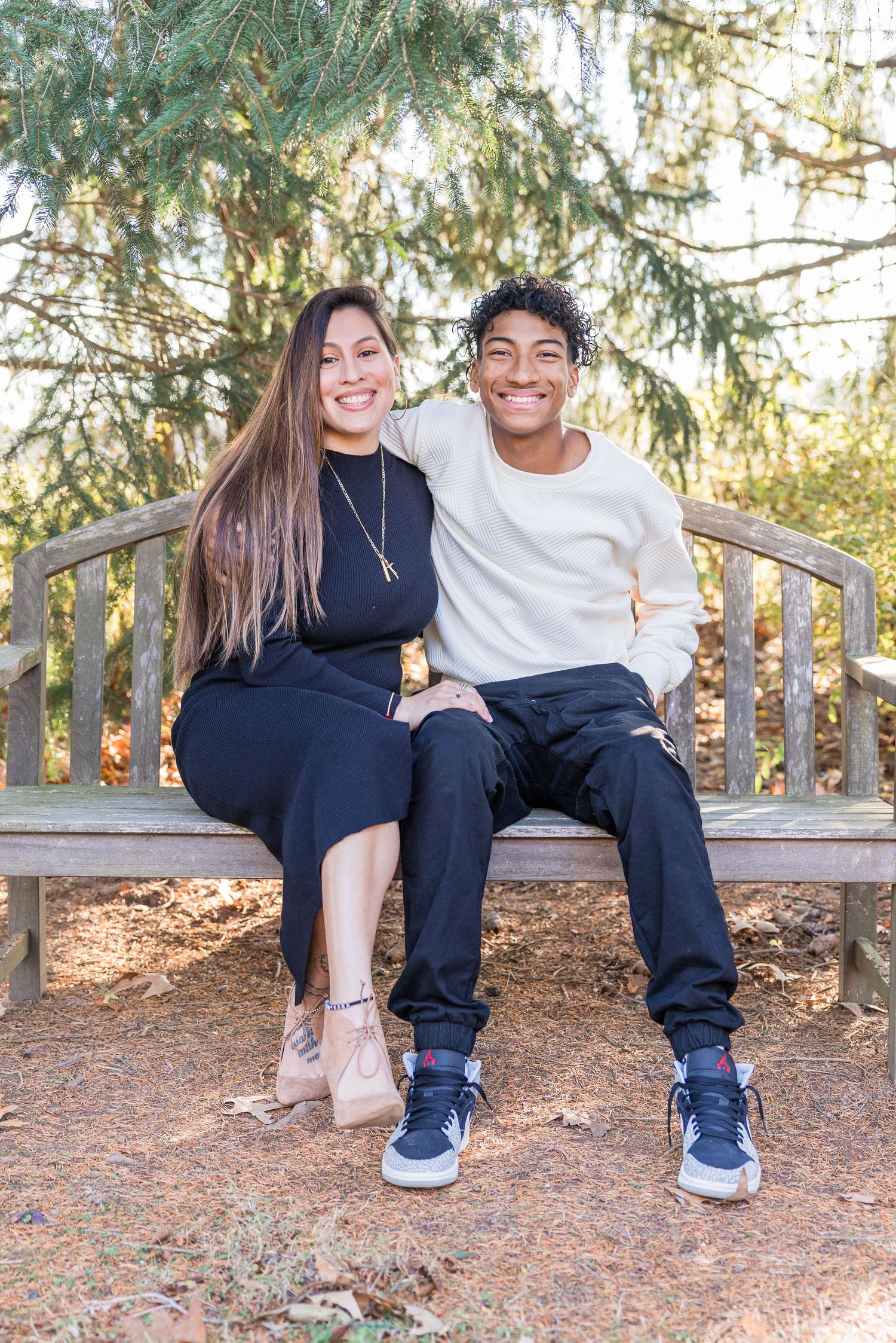 Two people sitting on a wooden bench surrounded by pine needles in a natural outdoor setting.