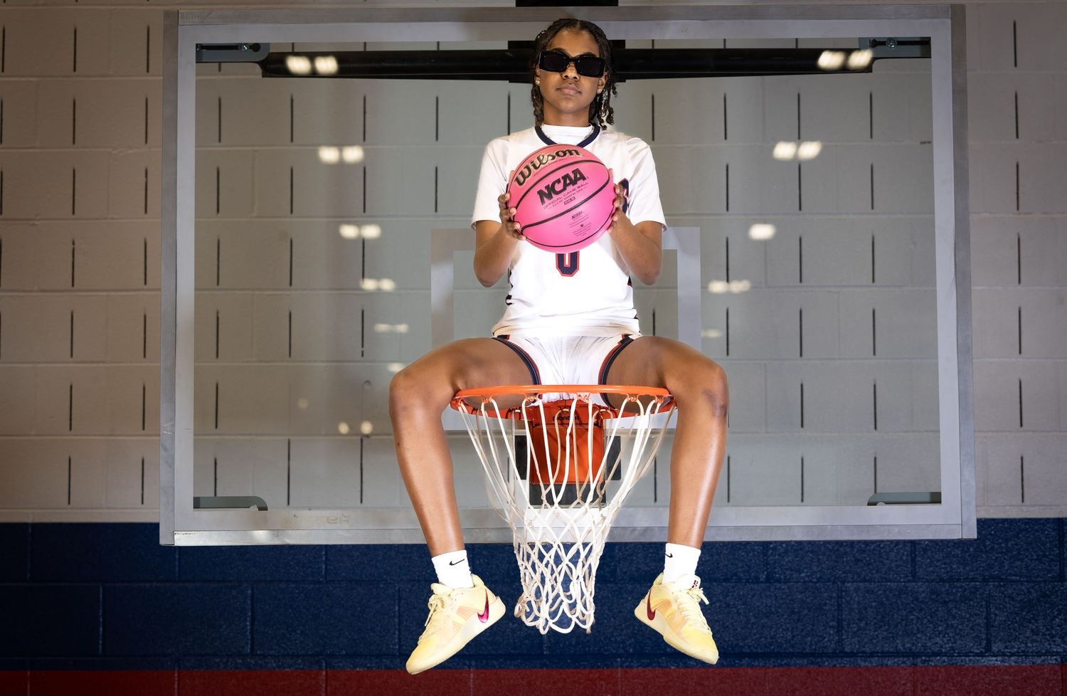 Athlete in white uniform sits on basketball hoop holding pink ball.