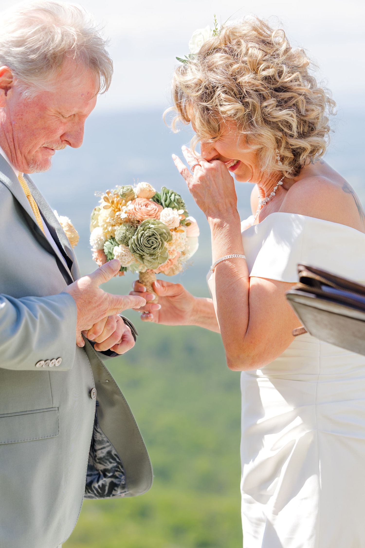 Older bride laughs emotionally as groom places wedding ring on her finger during outdoor mountain ceremony.