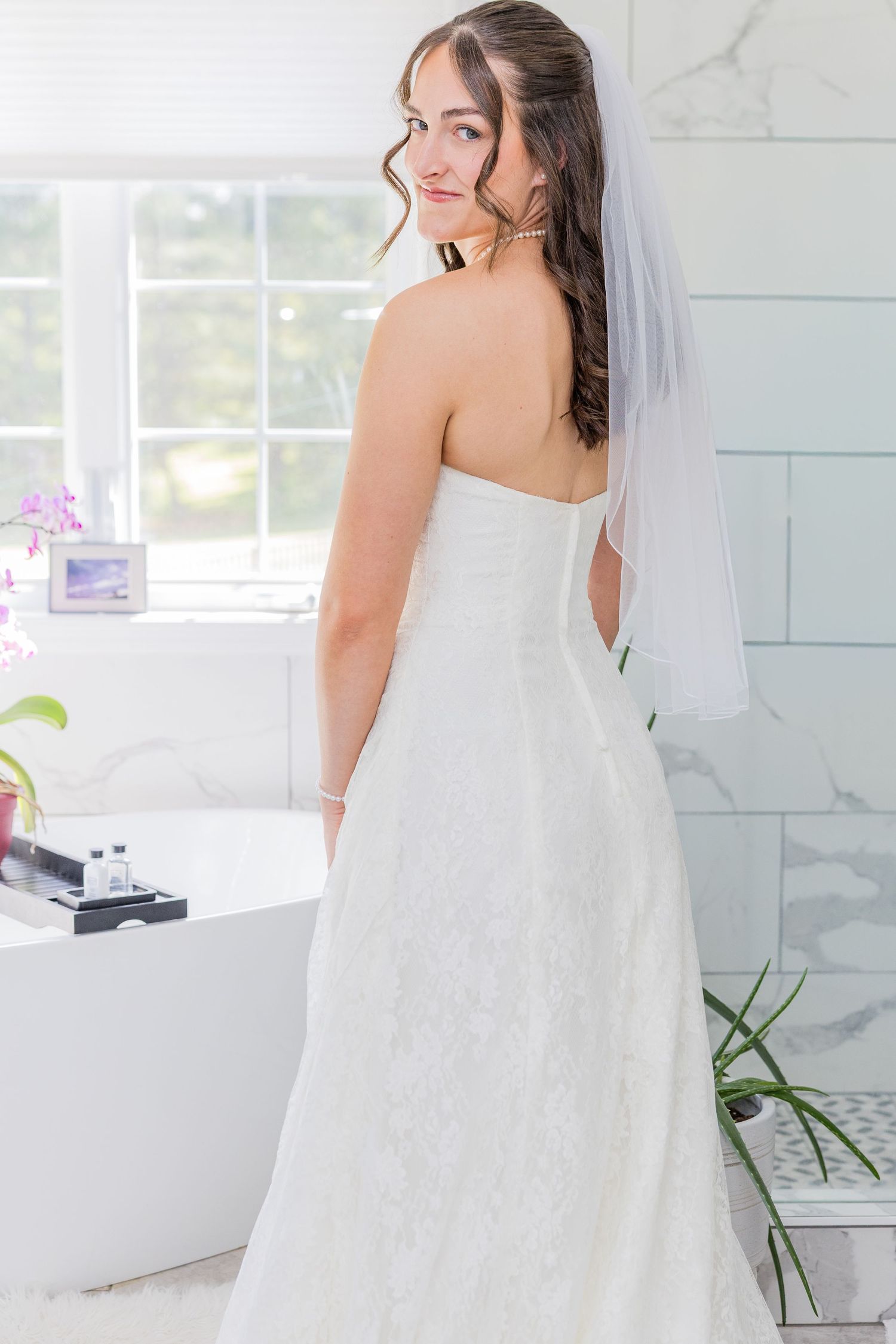 Bride in white strapless lace gown and veil poses in elegant bathroom, looking over shoulder toward camera.