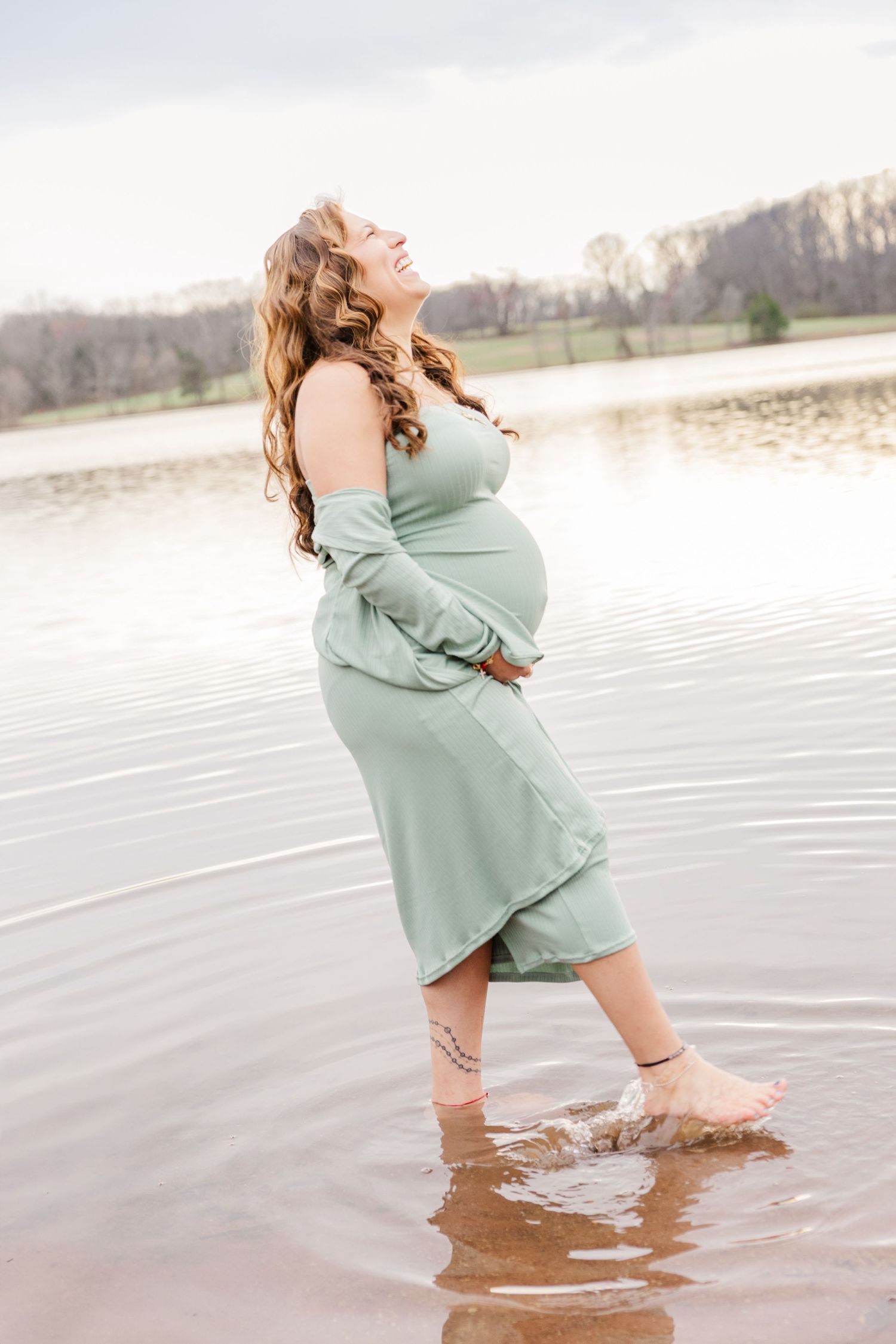 Artistic maternity photos of a woman in mint green dress wading in lake water during golden hour.