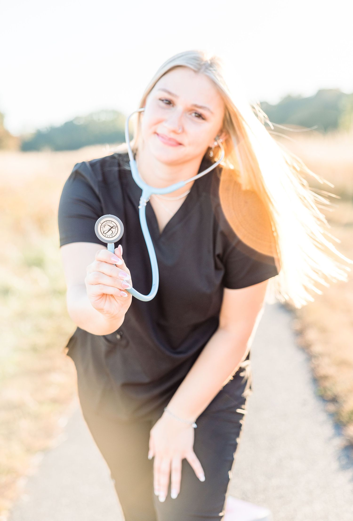Healthcare worker in black scrubs poses with stethoscope in golden sunlit outdoor setting.