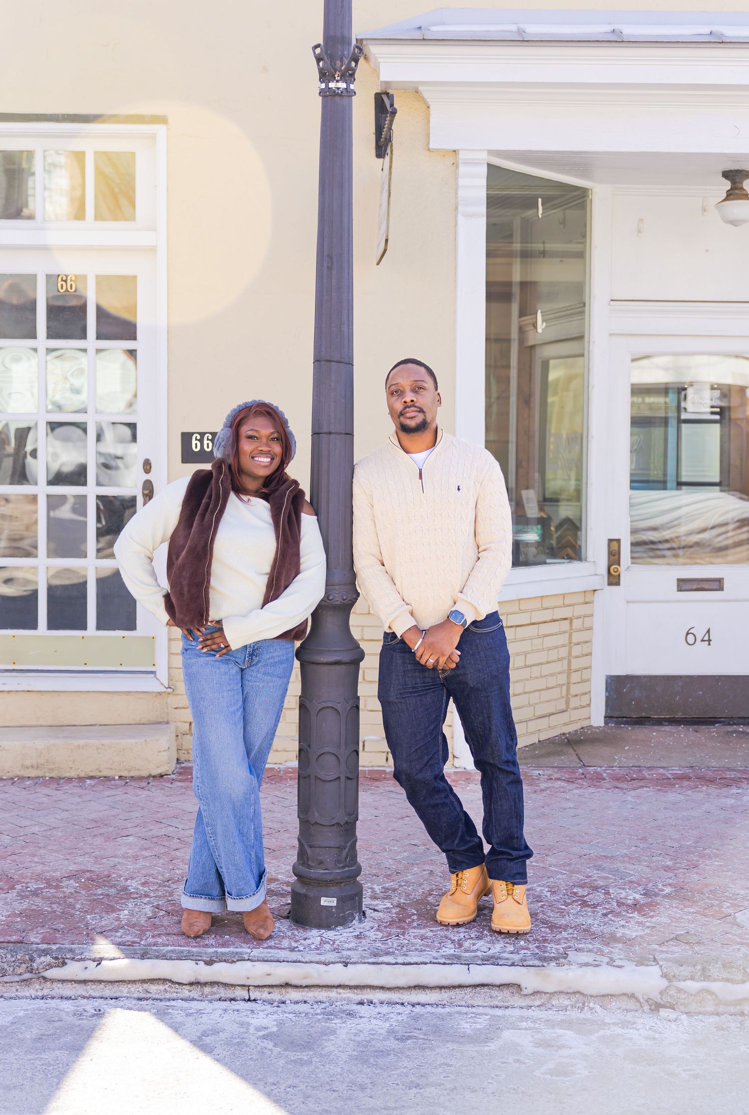 A couple stands by a lamp post on a snowy sidewalk in front of storefronts.