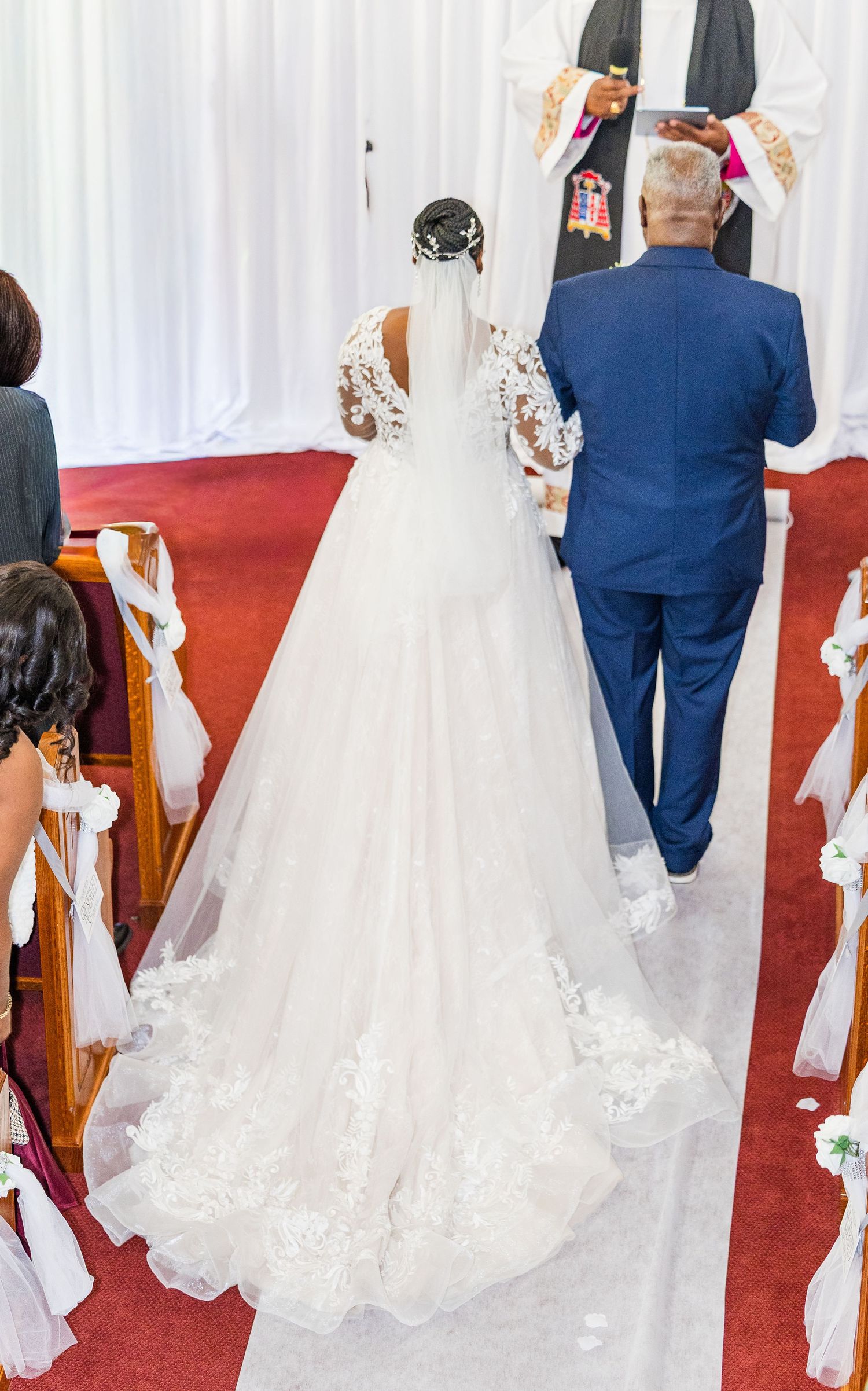 A bride in a flowing white lace gown with long train walks down a red-carpeted church aisle with someone in a blue suit.
